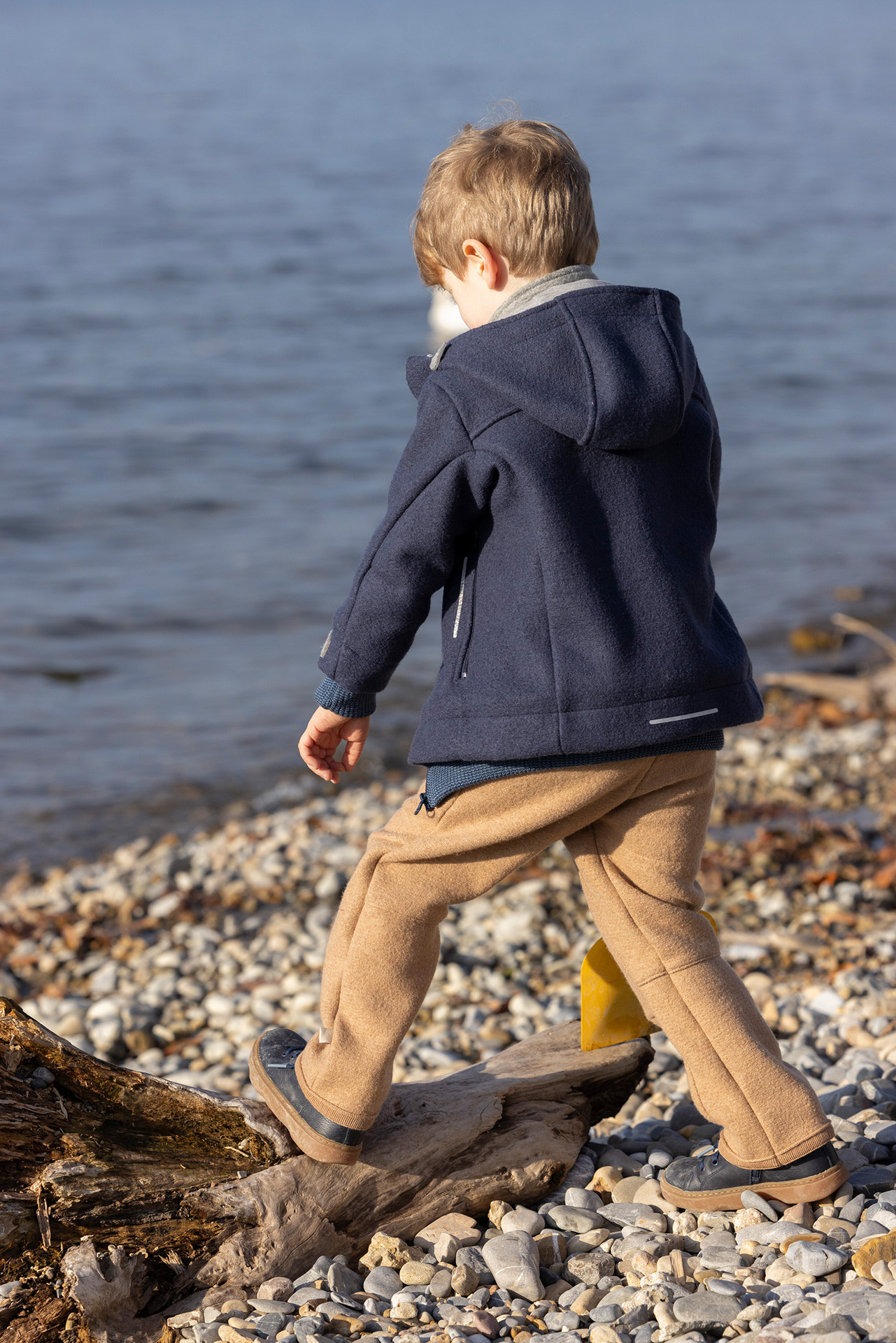 Junge in indigoblauer disana Outdoor-Jacke und karamellfarbener Walk-Hose klettert am steinigen Seeufer über Treibholz