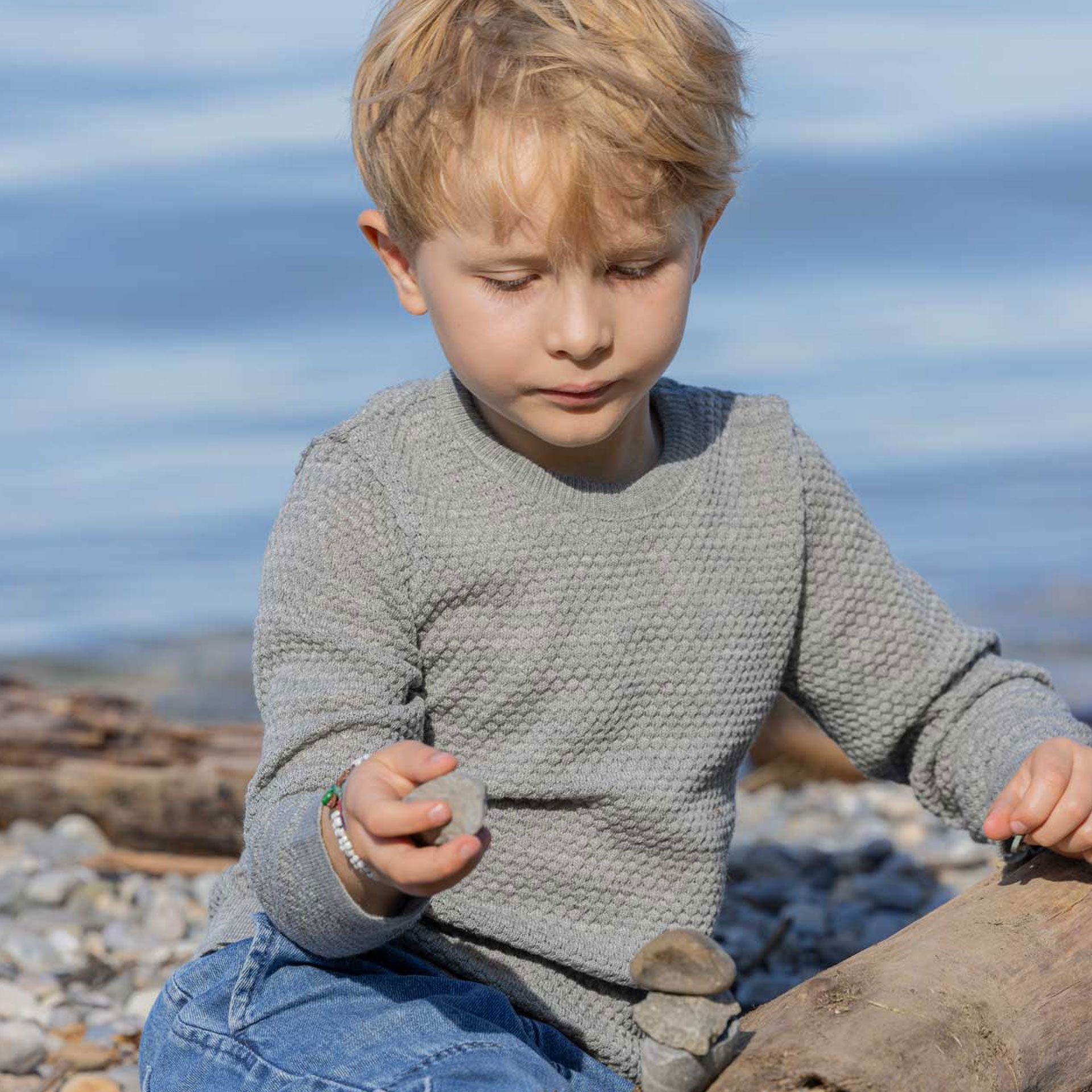 Blonder Junge im grauen disana Wabenstrick-Pullover aus Bio-Merinowolle beim Steine-Stapeln am Kieselstrand