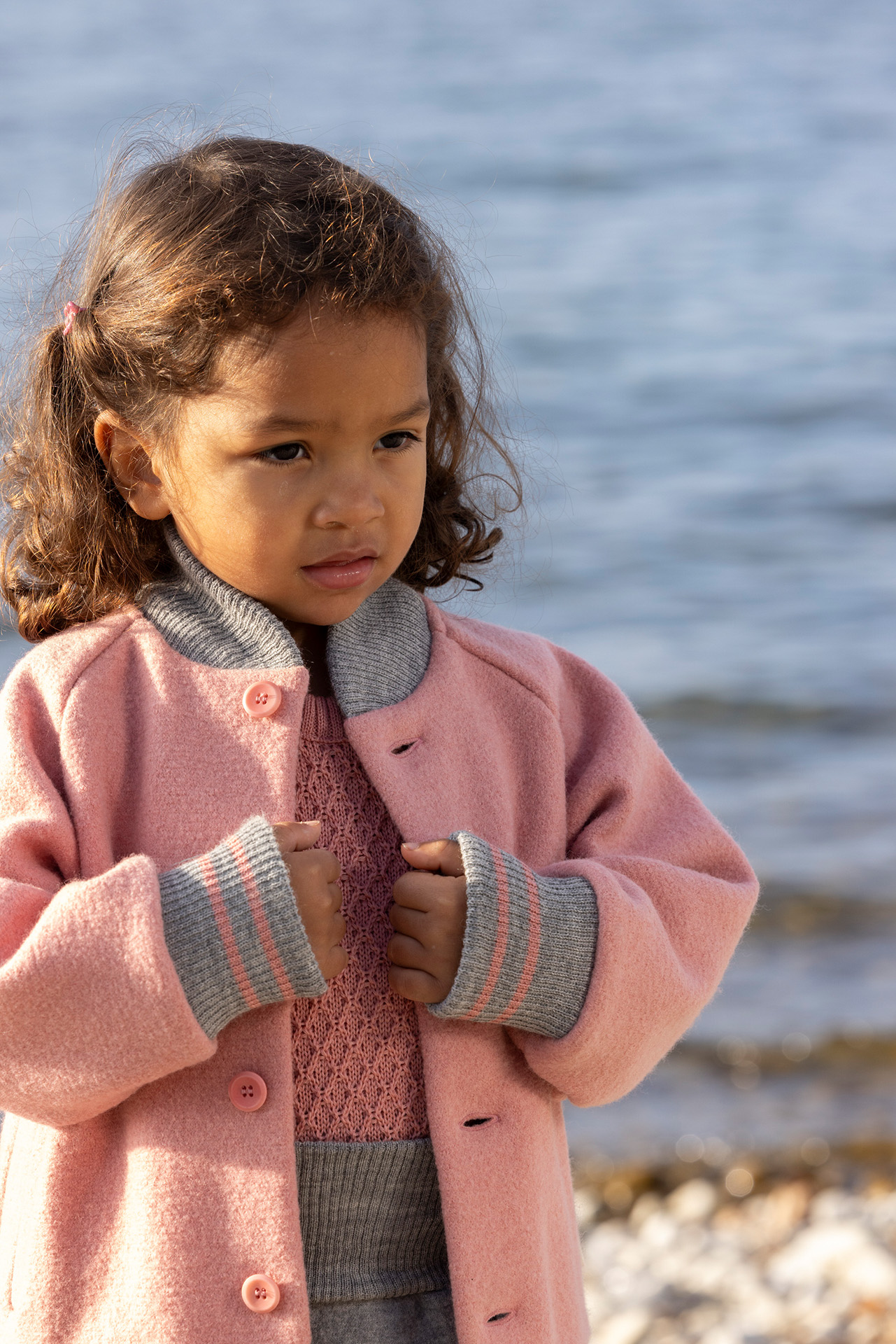 Mädchen mit  hellbrauner Haut in disana College-Jacke Softpink mit grau gestreiften Ärmelbündchen & Strickkragen Am Seeufer