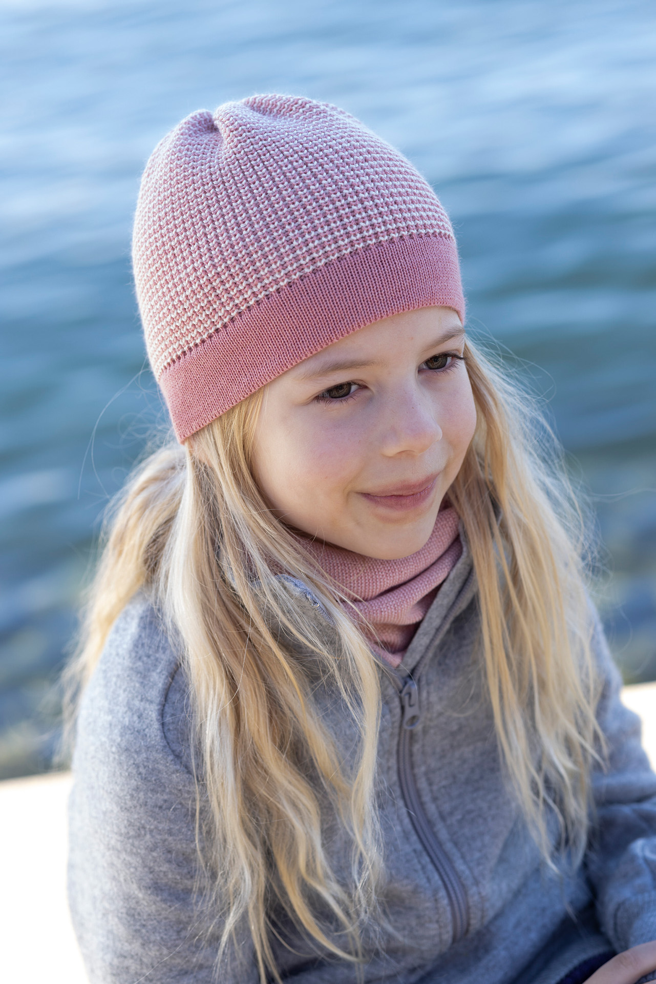 Blondes Mädchen mit langen Haaren trägt eine disana Beanie in Softpink-Natur und eine Zipper-Jacke in Grau vor Seehintergrund