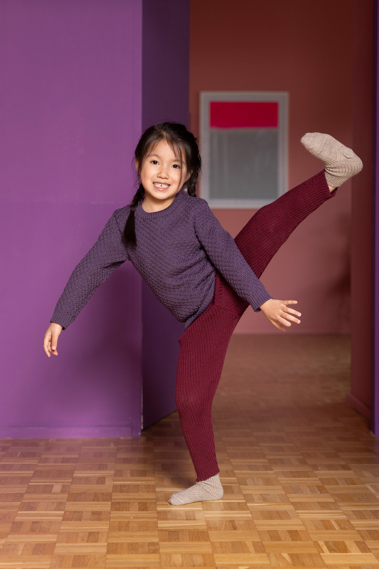 Asiatisches Mädchen führt ein Grand Battement aus in disana Aran-Pullover, Aubergine, Strick-Leggings Cassis auf Parkettboden