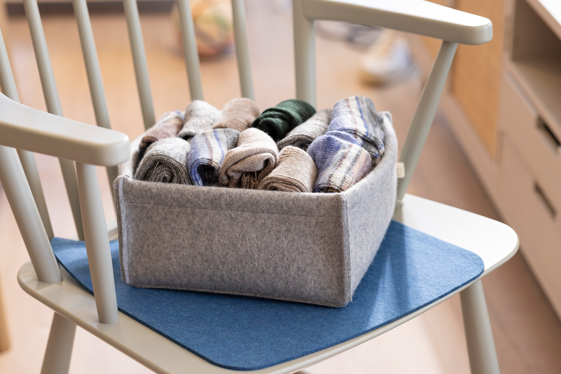 Graue disana Filz-Box mit eingerollten Wollsocken auf einer blauen Filz-Auflage und einem Holzstuhl. Filz aus Bio-Merinowolle