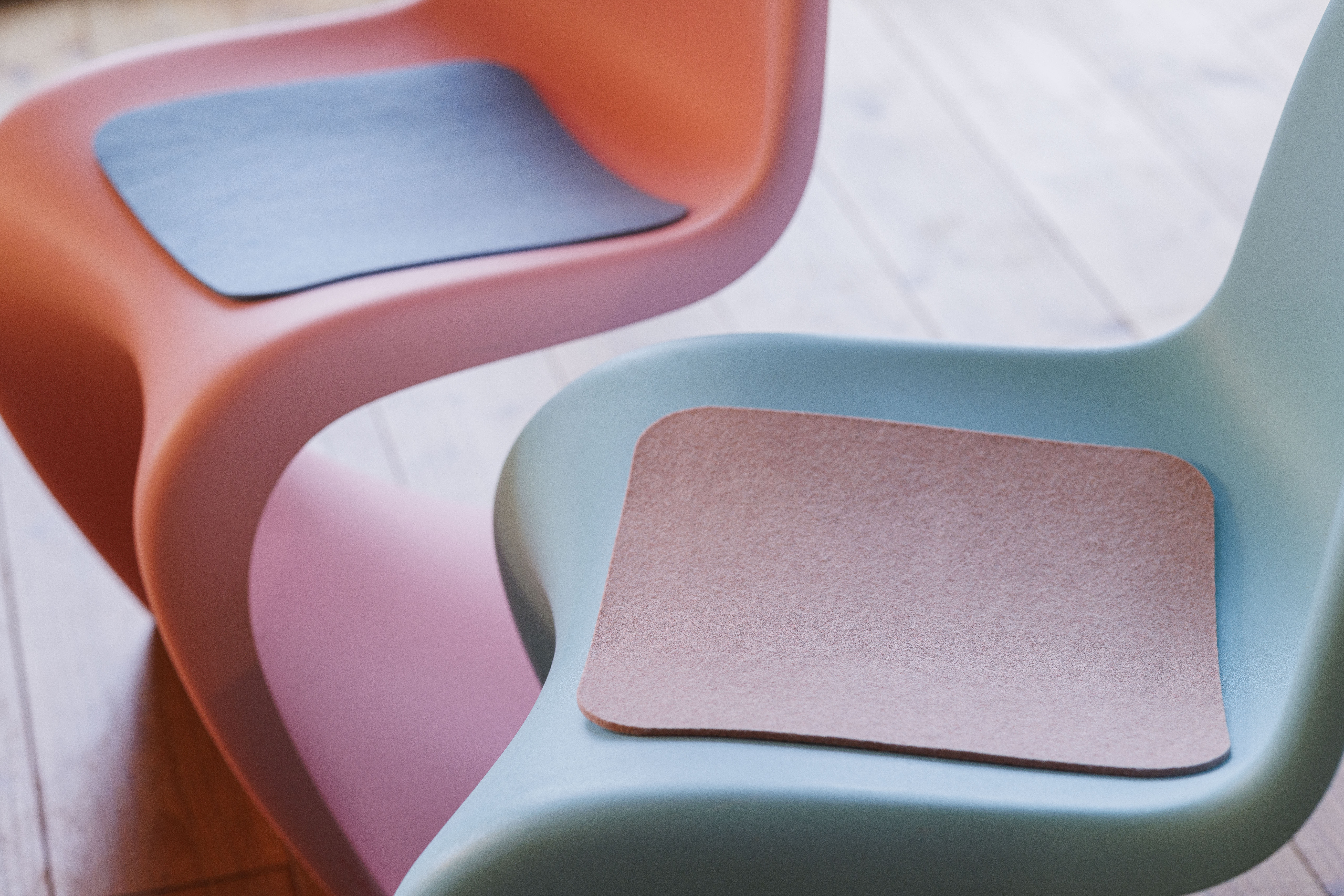 Zwei disana Filz-Auflagen in Rosé und Lagoon auf modernen Design-Stühlen in Orange und Hellblau.