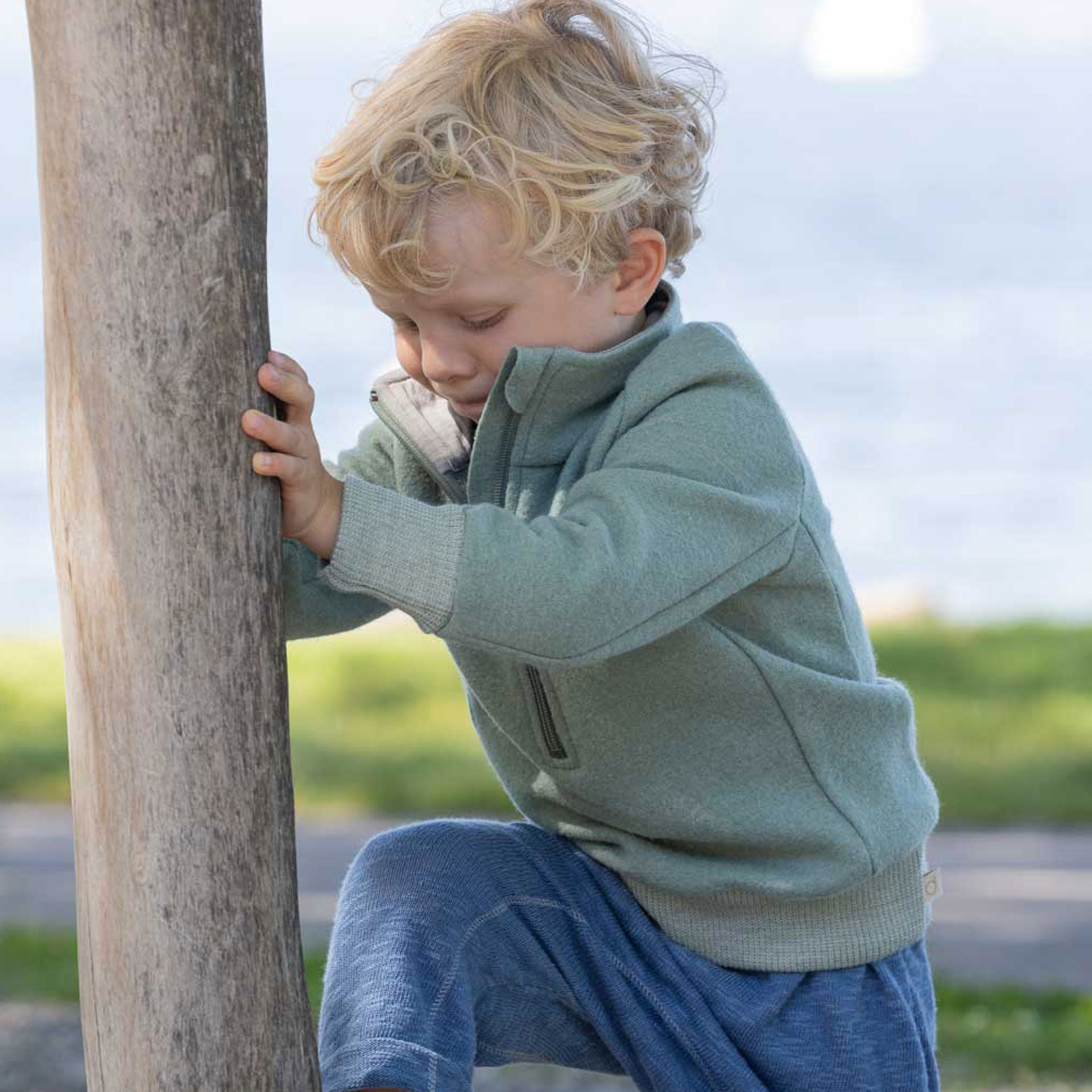 Blondes Kleinkind mit jadefarbenem Troyer und Leinen-Strickhose Taubenblau klettert auf einen Baumstamm auf dem Spielplatz.