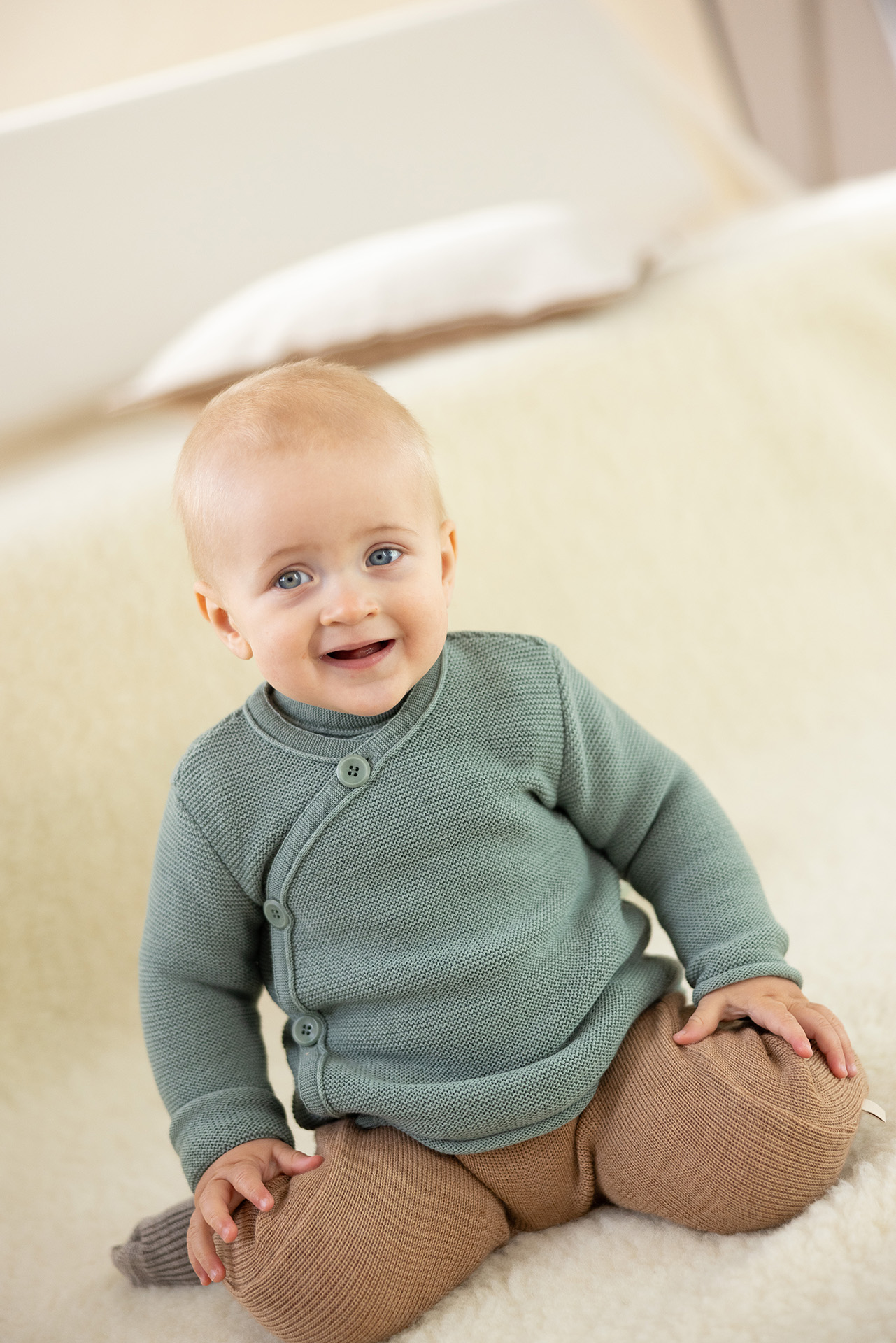 Blondes Baby mit blauen Augen in Babyjacke Jade & Strick-Trägerhose in Karamell sitzt mit angewinkelten Beinen auf Schafffell
