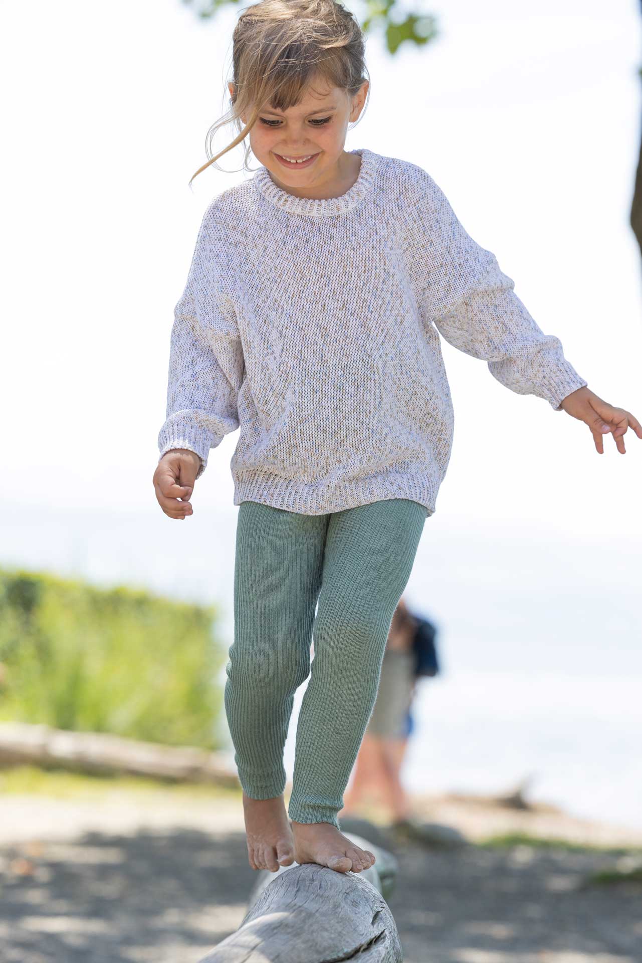 Dunkelblondes Mädchen in disana Baumwoll-Pullover "Spray Natur" & Leggings in Jade balanciert barfuß auf Holzstamm im Freien