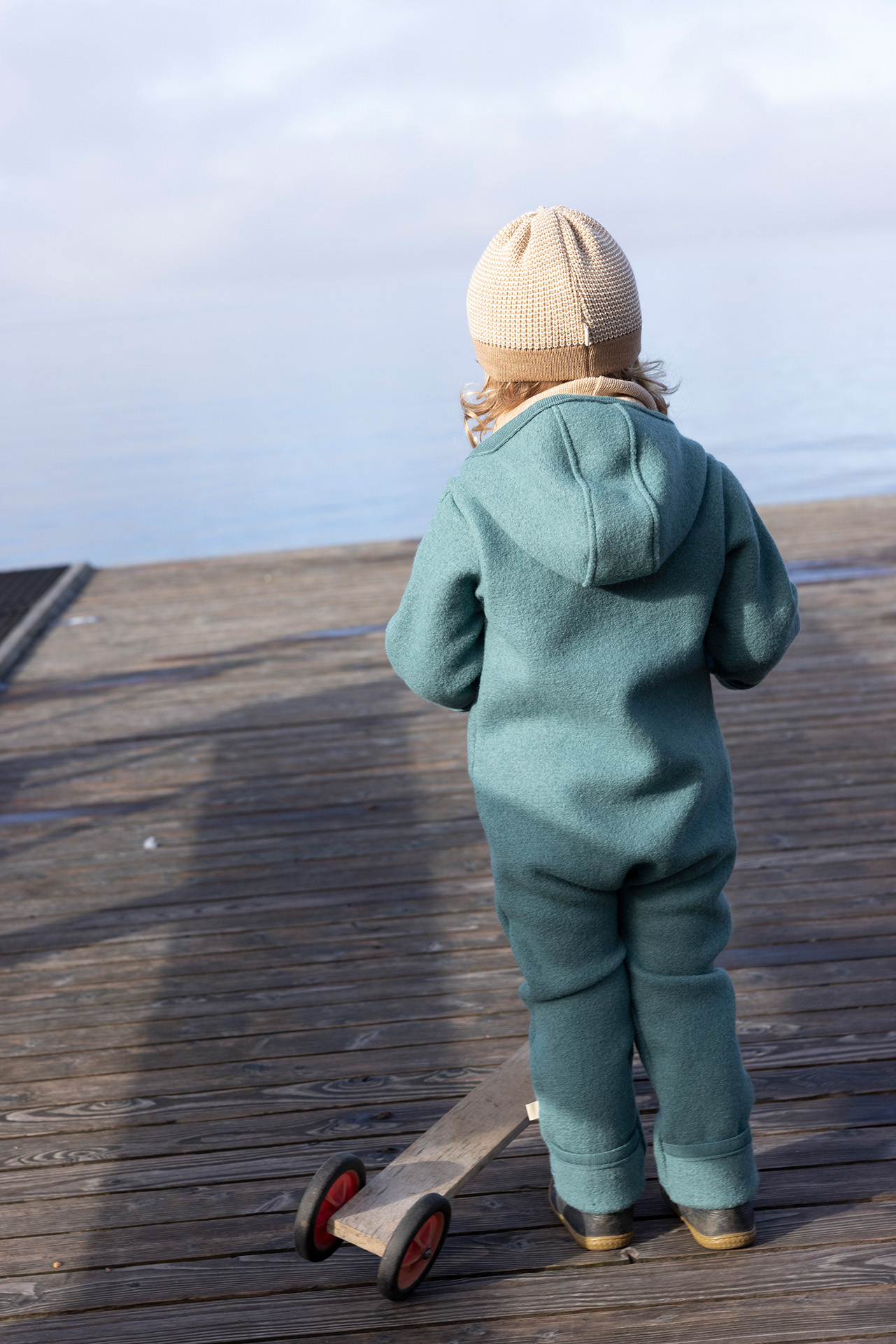Rückansicht eines Kindes in disana Walk-Overall Eukalyptus und Beanie in Karamell-Natur mit Holzroller auf einem Steg am See