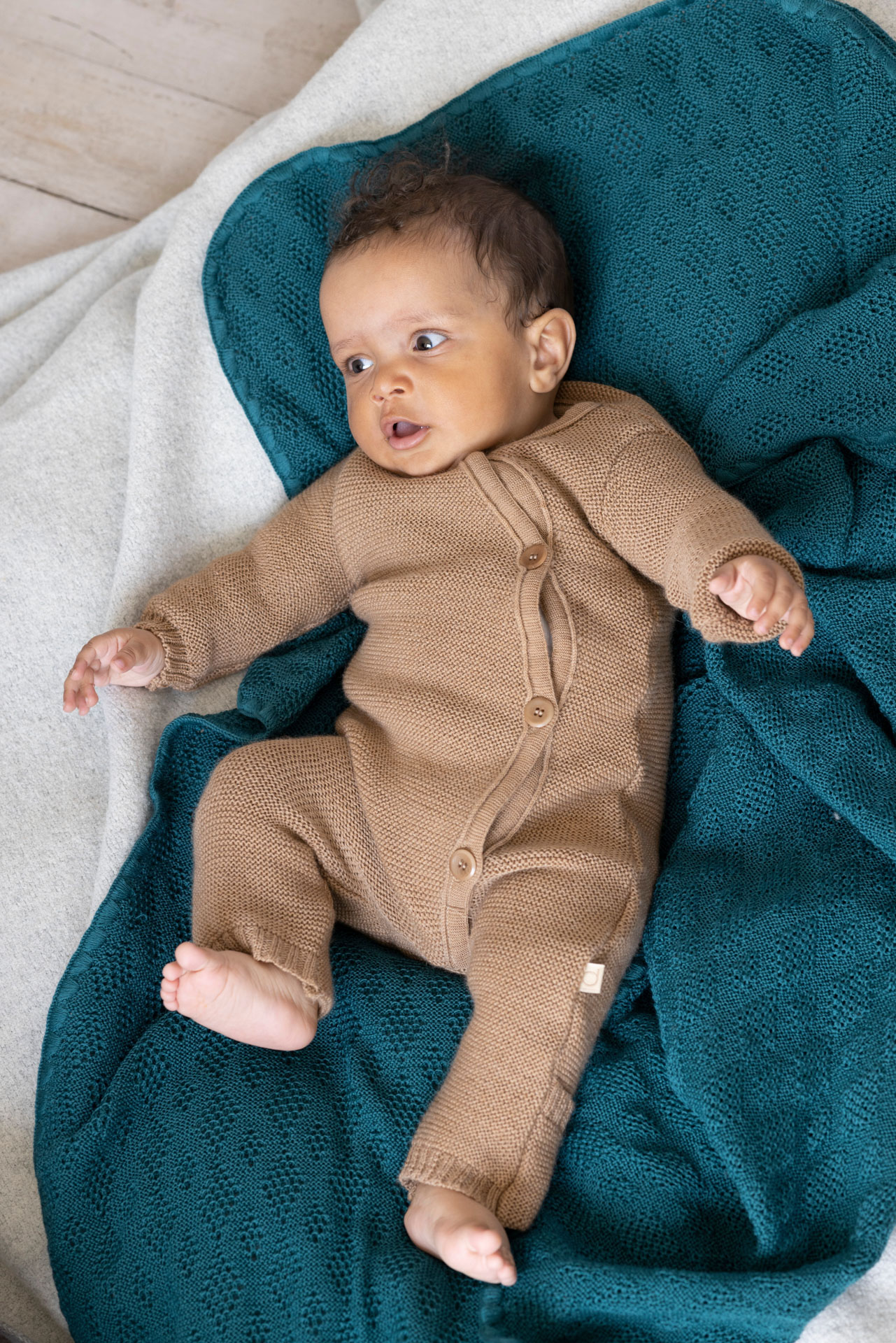Baby in disana Strick-Overall, Karamell liegt auf einer Woll-Babydecke in Pacific mit Ajour-Muster.