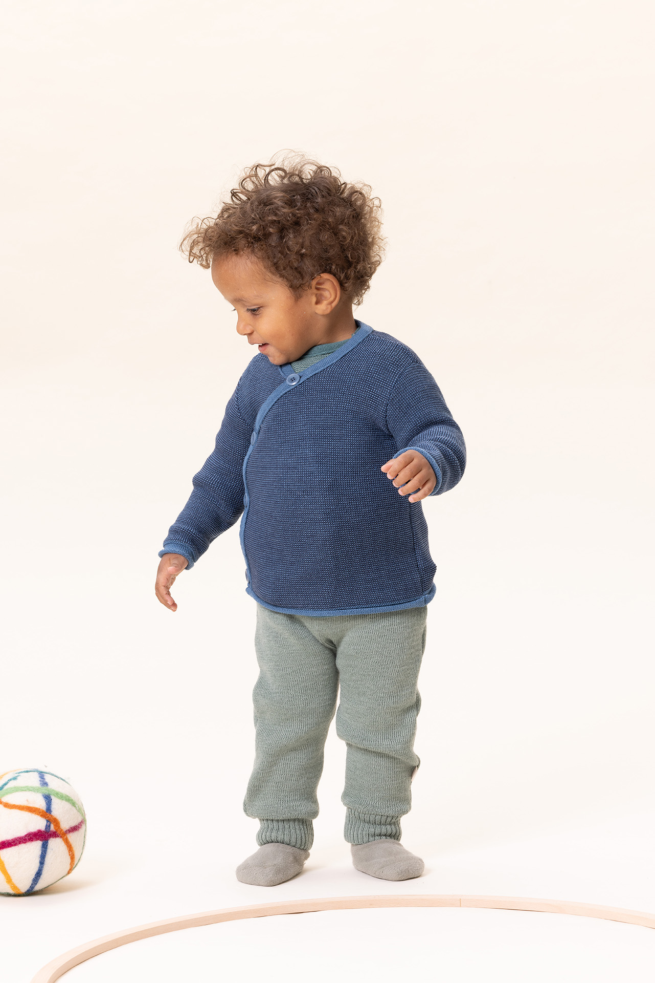 Kleinkind mit hellbrauner Haut in disana Melange-Jacke Taubenblau-Indigo & Strick-Trägerhose Jade beim Spielen mit Filzball
