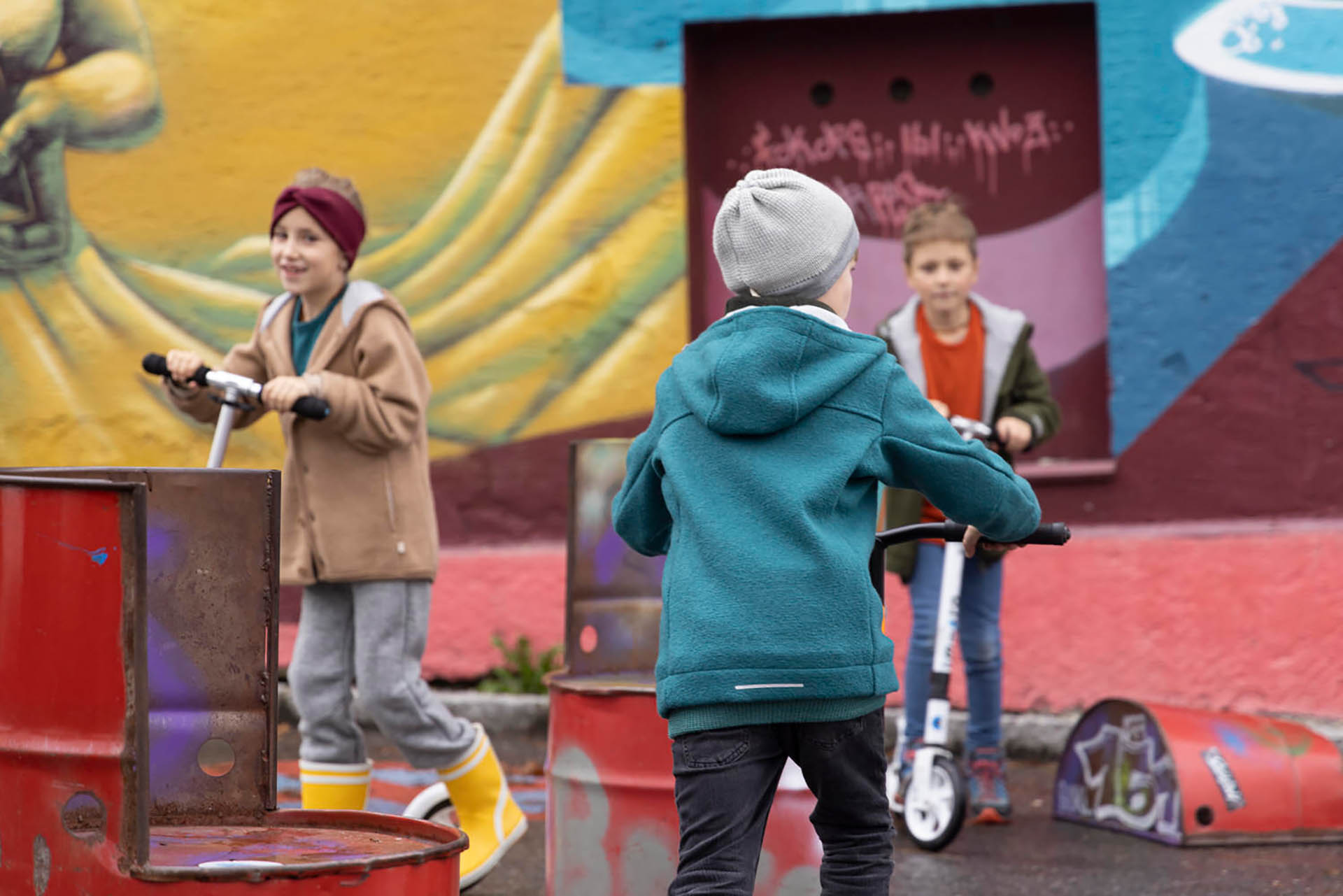 Mehrere Kinder in disana Wollwalk-Kleidung aus Bio-Merinowolle spielen mit Rollern vor einer bunten Graffiti-Wand