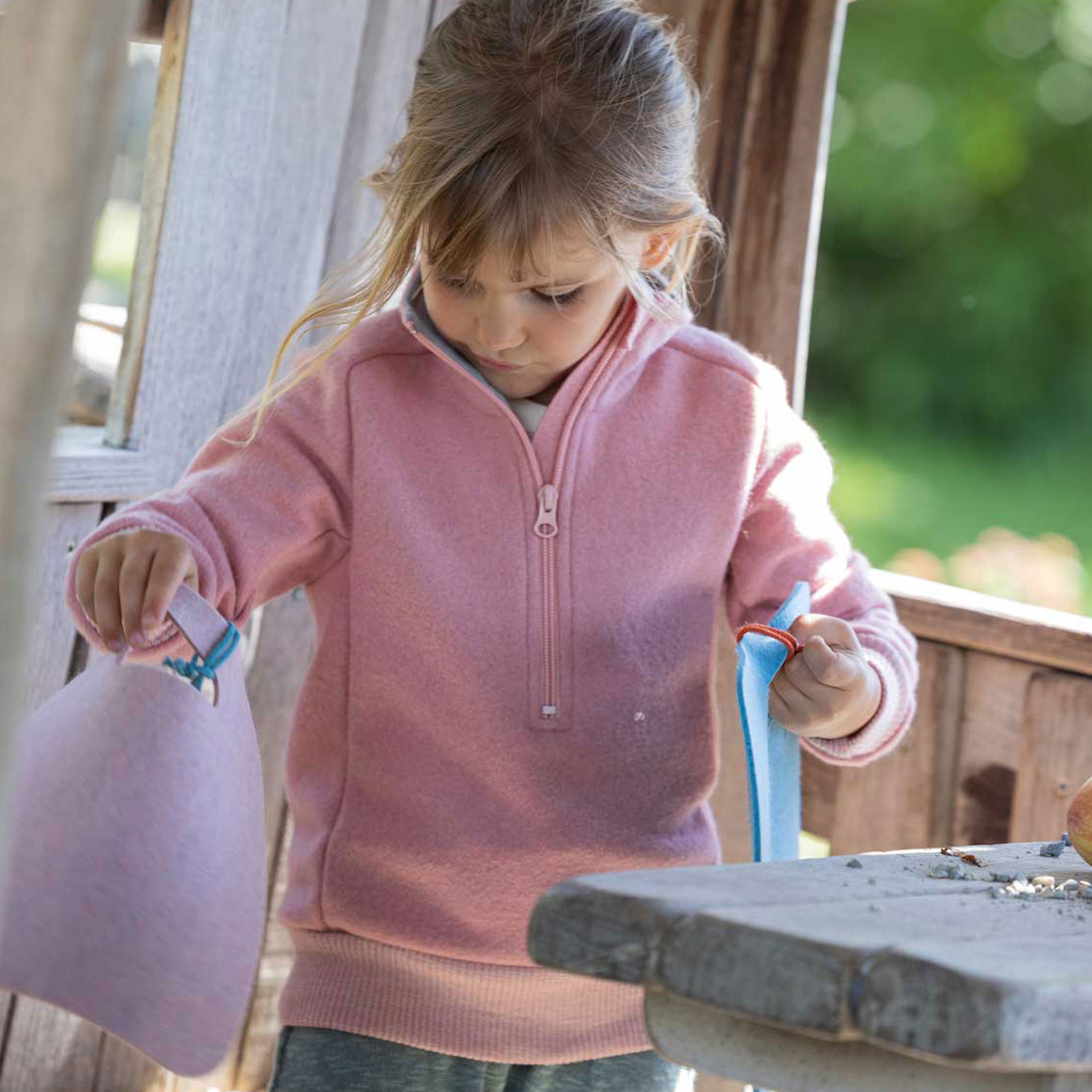 Mädchen im softpinkfarbenen disana Troyer aus Bio-Merinowolle beim Spielen an einem Holztisch im Kletterhaus