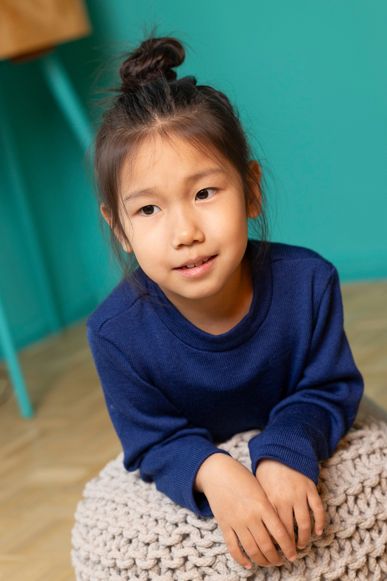 Asiatisches Mädchen in disana indigofarbenem Strick-Pullover vor einer grünen Wand, gestützt auf einen hellen Strick-Puff
