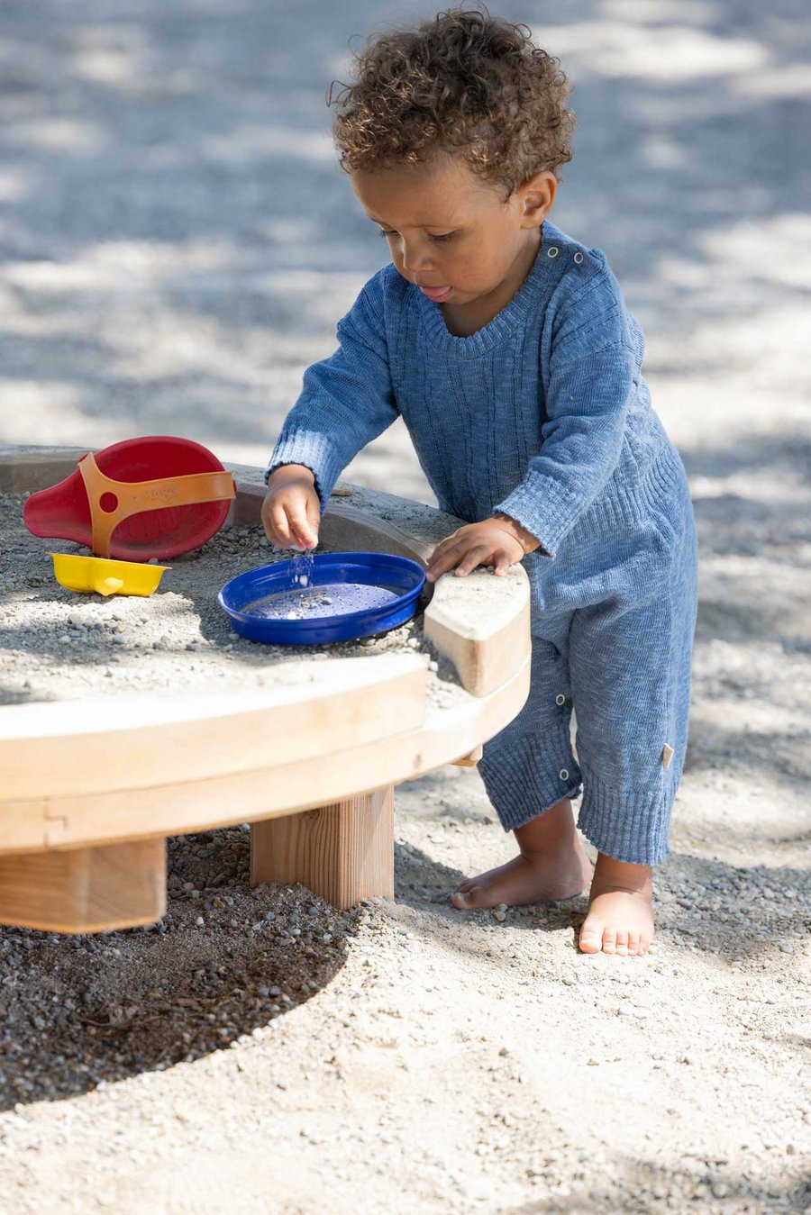Dunkelhäutiges Kleinkind trägt disana Leinen-Strampler, Taubenblau & spielt barfuss im Sandkasten mit einem blauen Teller