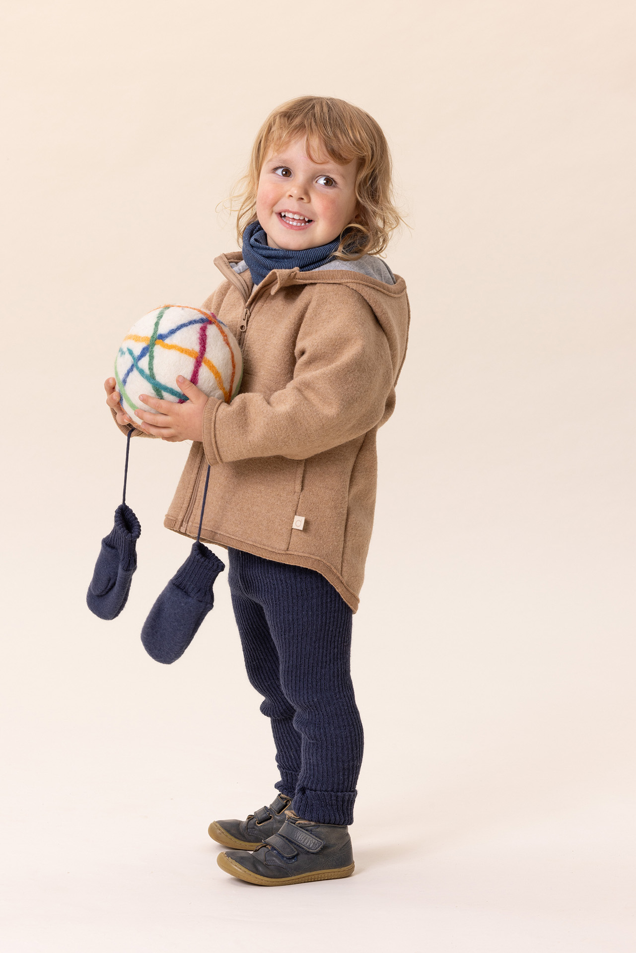 Kind in disana Walk-Jacke Karamell, Strick-Leggings in Indigo hält einen Filzball & blickt in die Kamera - beiger Hintergrund