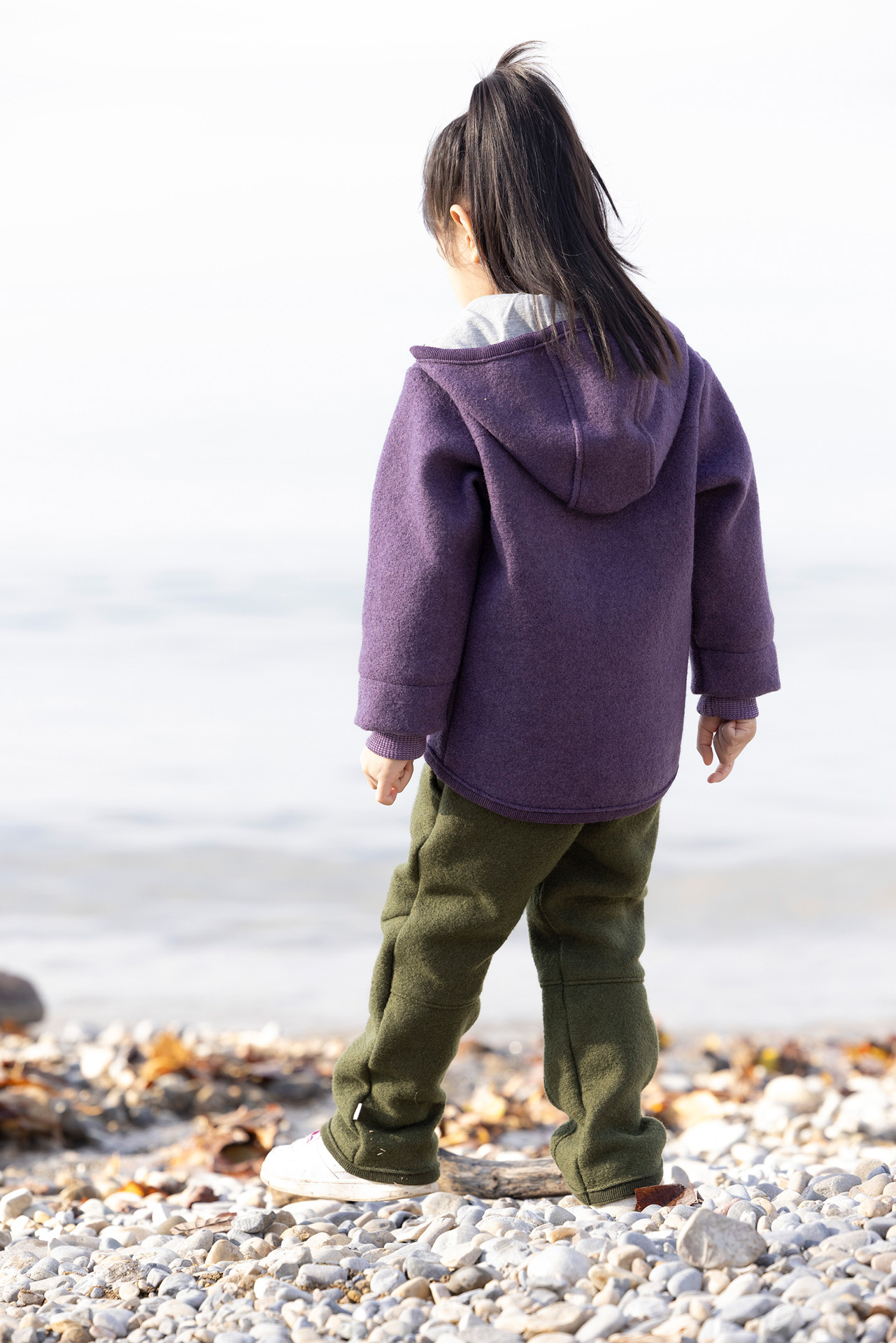 Rückansicht eines asiatischen Mädchen in disana Walk-Jacke Aubergine & Walk-Hose in Oliv am Kiesstrand eines Seeufers