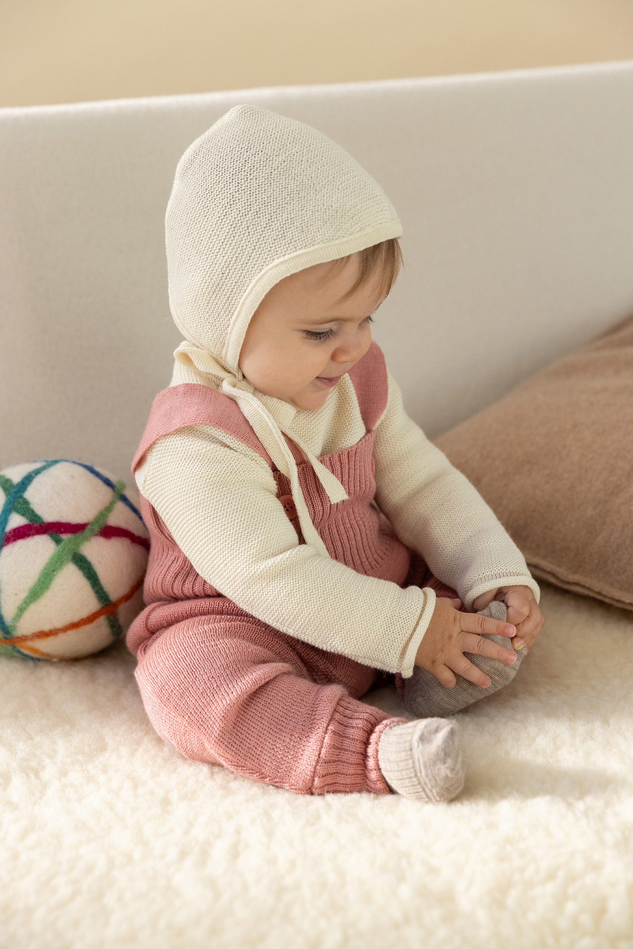 Baby in disana Strick-Trägerhose Softpink, Baby-Pullover & Strick-Häubchen, Natur hält Fuß auf hellem Lammfell sitzend