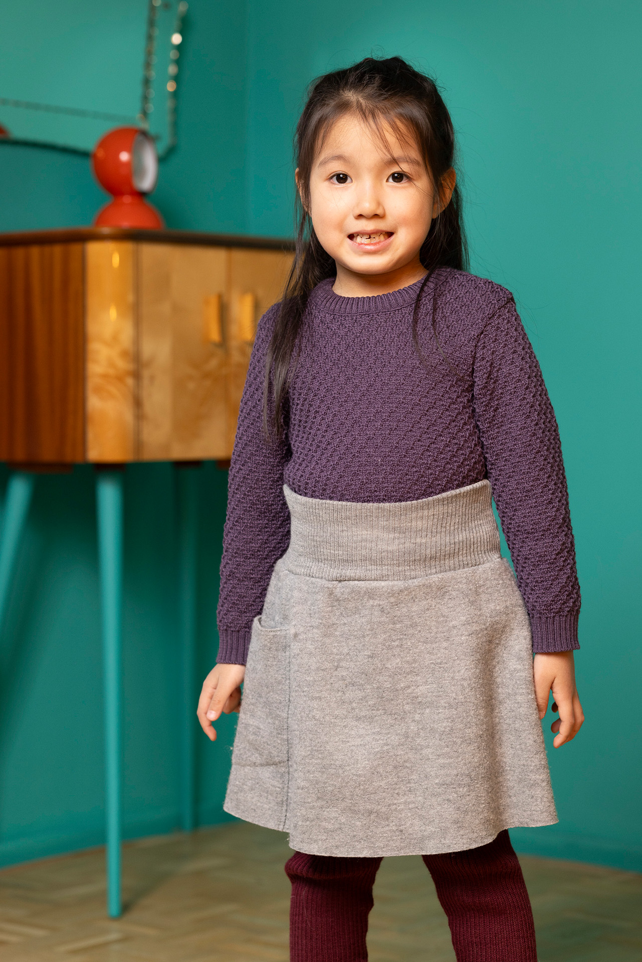 Asiatisches Mädchen in Aran-Pullover, Aubergine, Walk-Rock, Grau & Strick-Leggings Cassis, stehend vor grünem Hintergrund.
