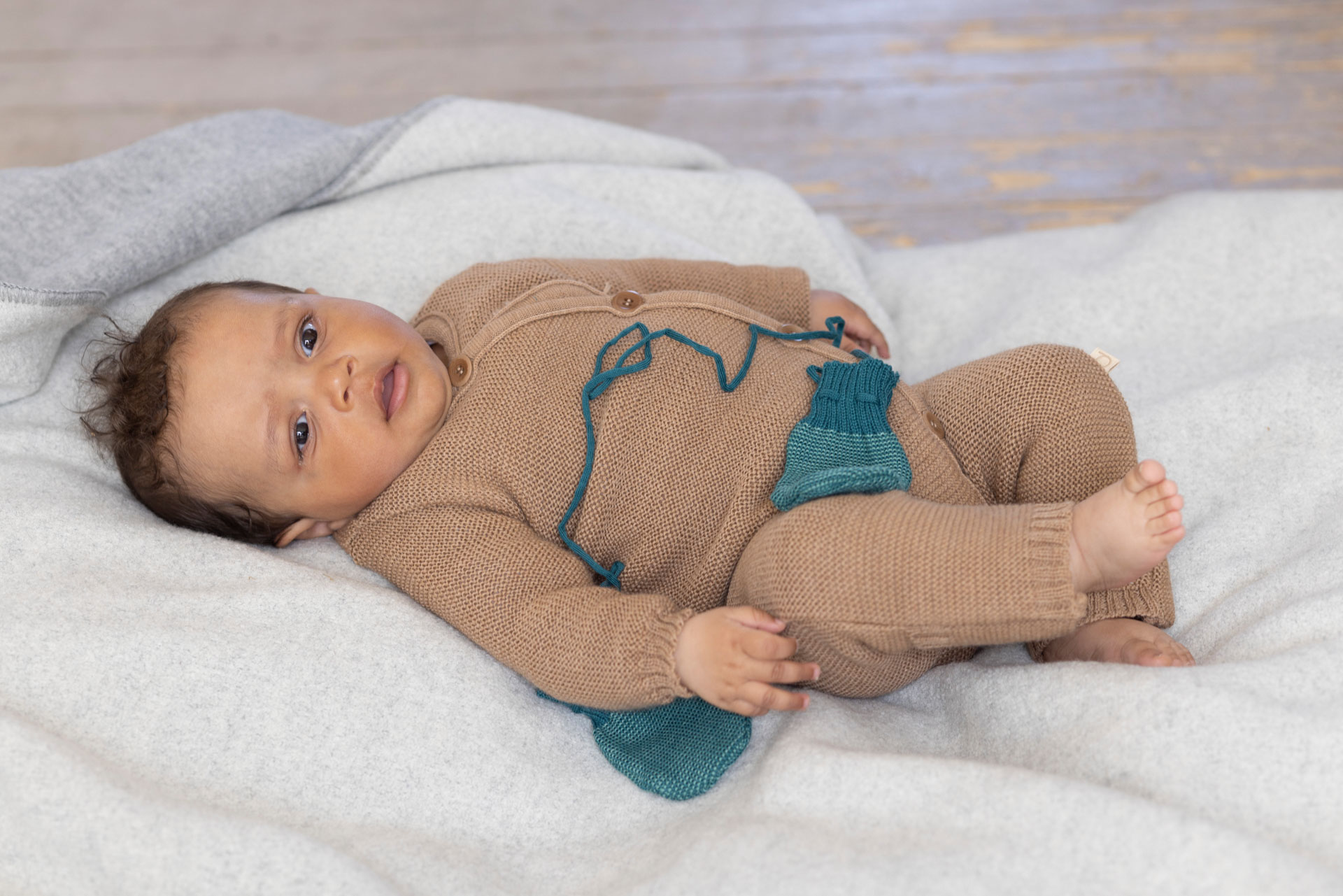 Baby in brauner Strickkleidung liegt auf einer hellen Wolldecke