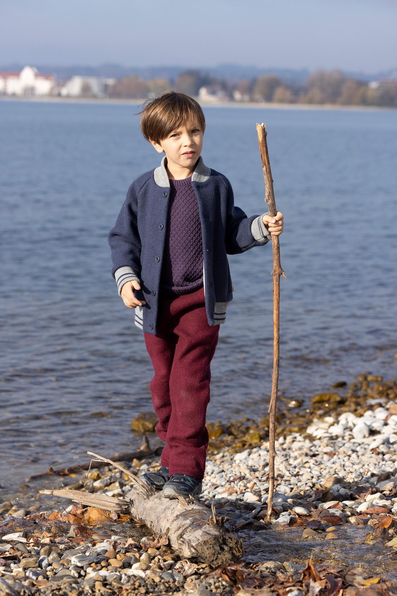 Junge, braune Haare in disana College-Jacke, Indigo, Walkhose in Cassis balanciert mit Stock auf einem Holzstamm am Seeufer
