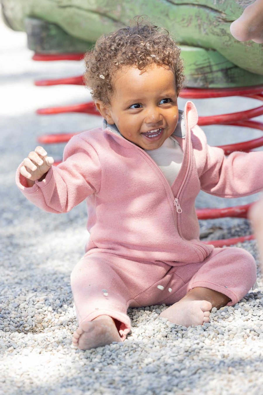 Dunkelhäutiges Kind im disana Walk-Anzug Softpink sitzt auf Kieselsteine & lacht, Sommer-Wollwalk 260g/m² aus Bio-Merinowolle