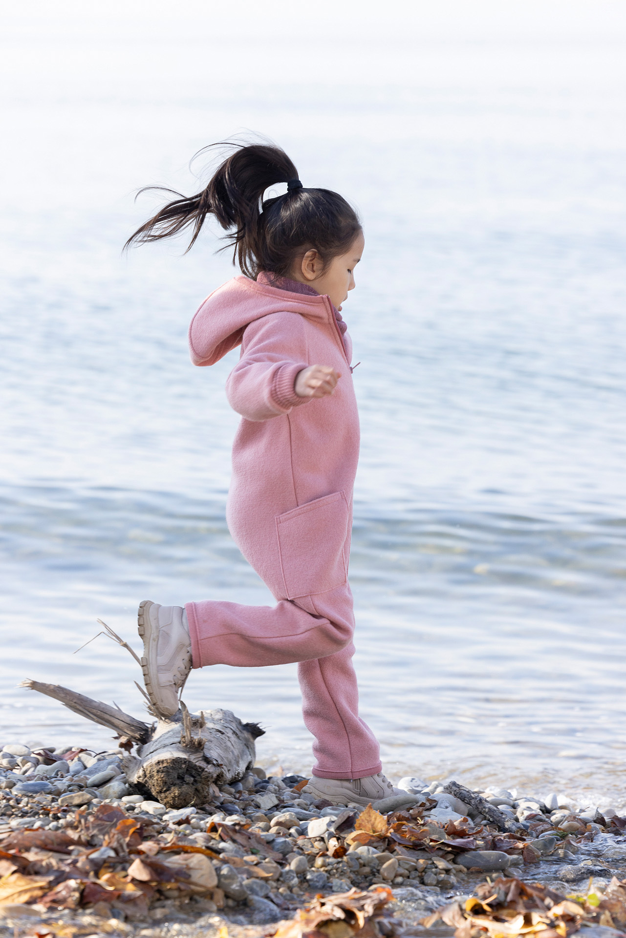 Asiatisches Mädchen im disana Walk-Overall Softpink und Schlauch-Schal in Cassis-Rosé läuft am Kieselstrand am Seeufer.