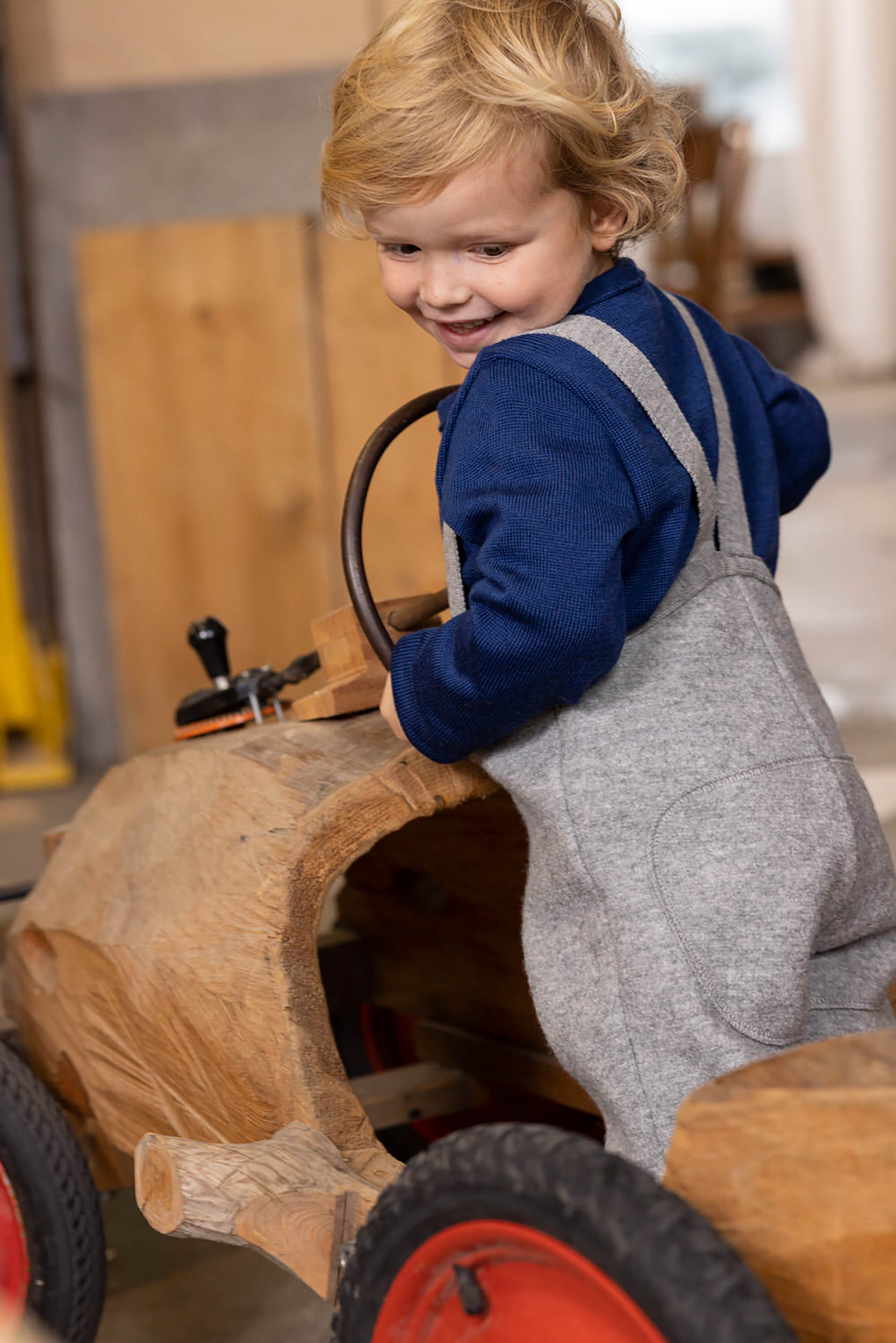 Blondes Kind in disana Walk-Hose in Grau mit Trägern und Strick-Pullover in Marine spielt auf einem Holz-Traktor
