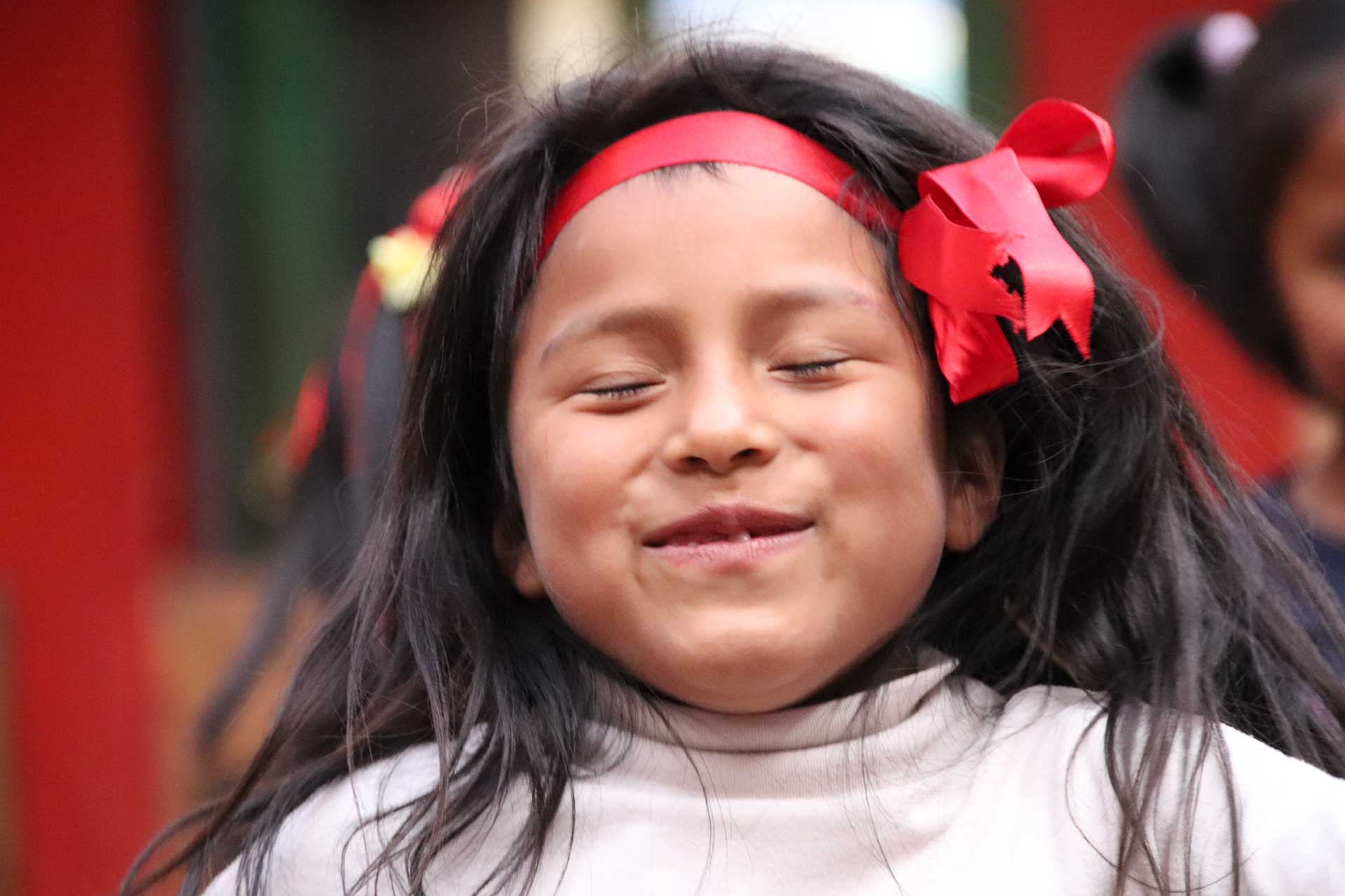 Casa Mantay, Cusco, Peru: Lächelndes Mädchen mit geschlossenen Augen, dunklen Haaren & einem roten Haarband mit Schleife.