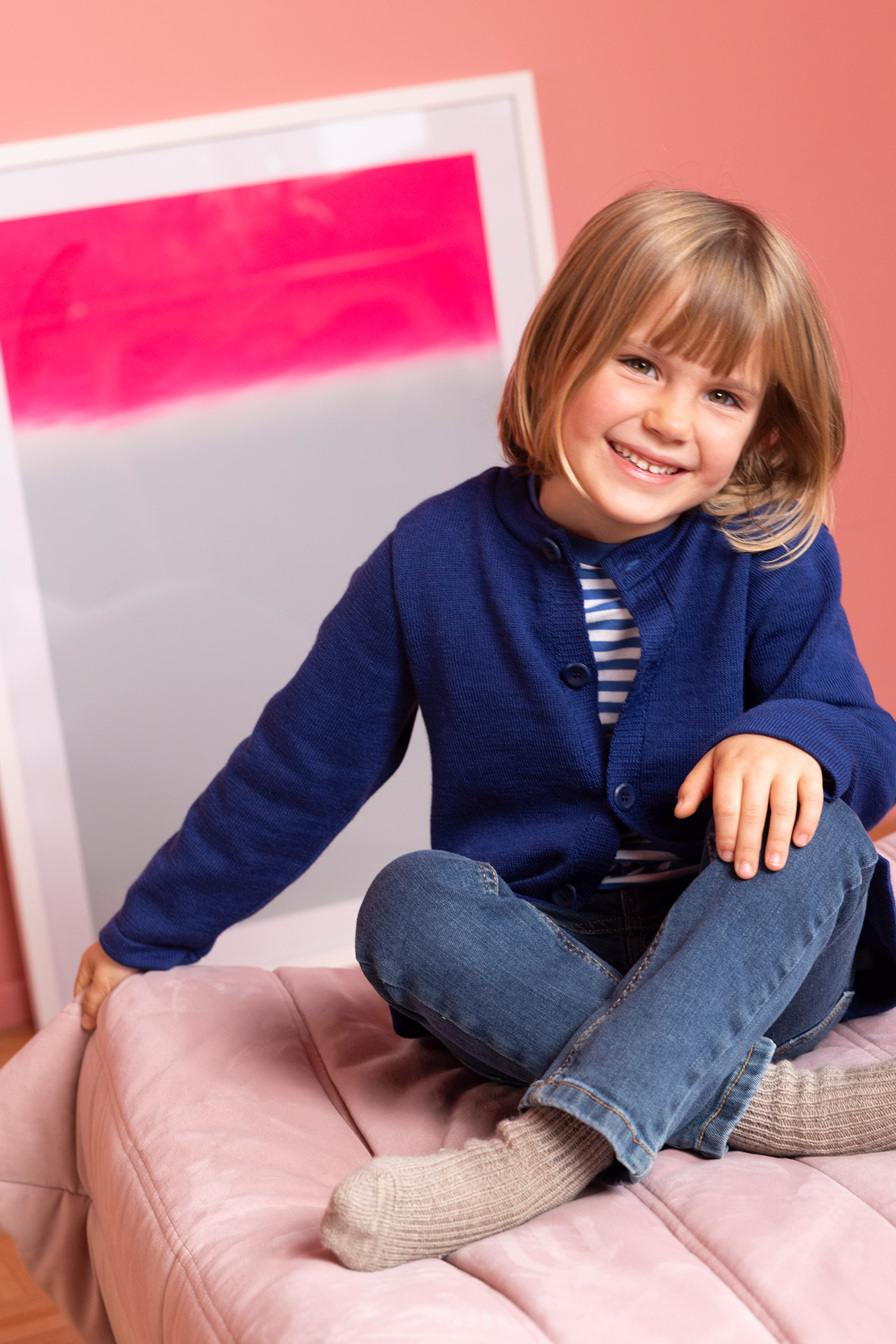 Blondes lachendes Kind in disana Strick-Jacke Indigo & Jeans sitzt auf rosa Polster im Hintergrund ein Bild mit pinkem Akzent