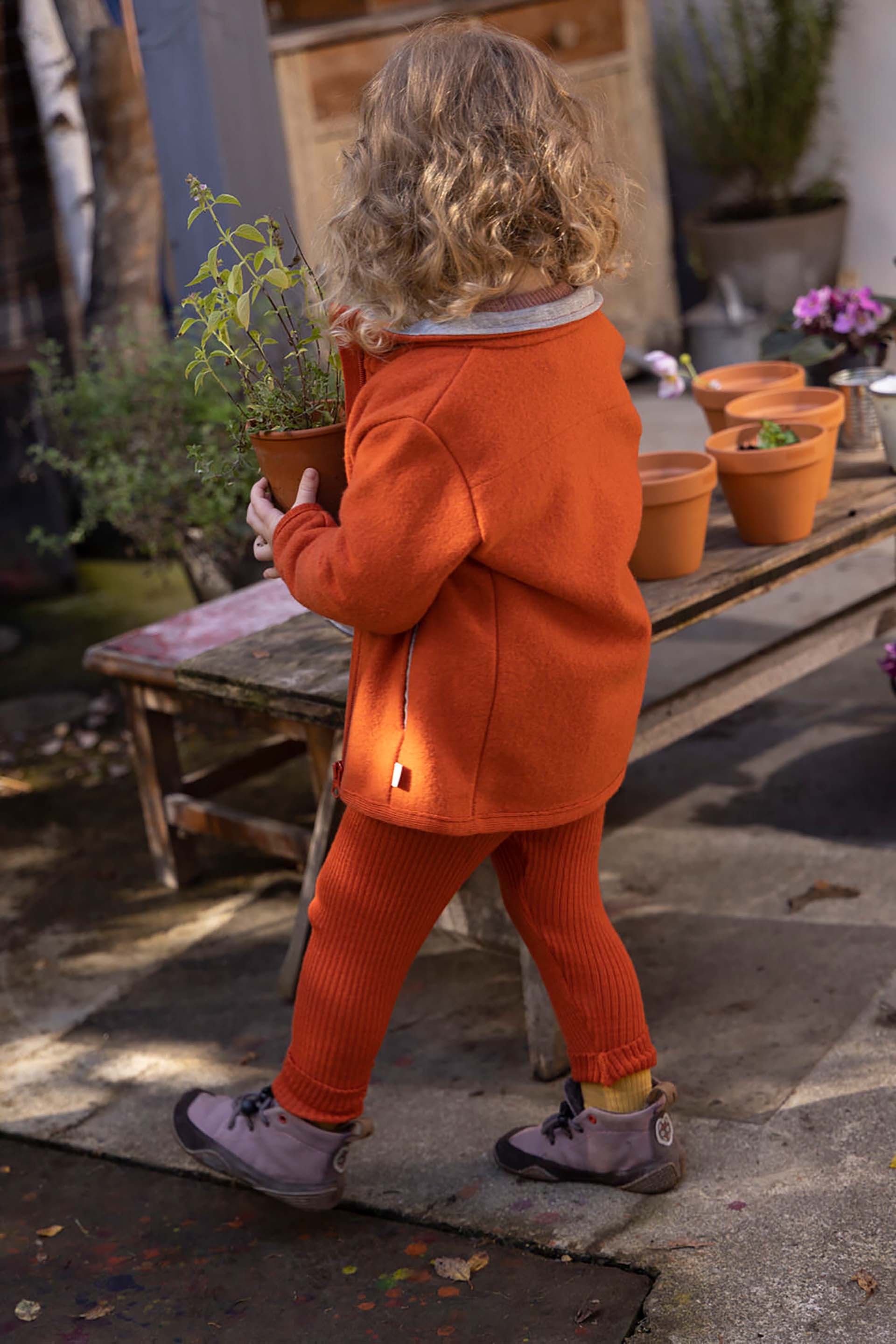 Rückansicht eines blonden Kindes in disana Zipper-Jacke und Leggings in Orange in einem Hof mit mehreren Blumentöpfen