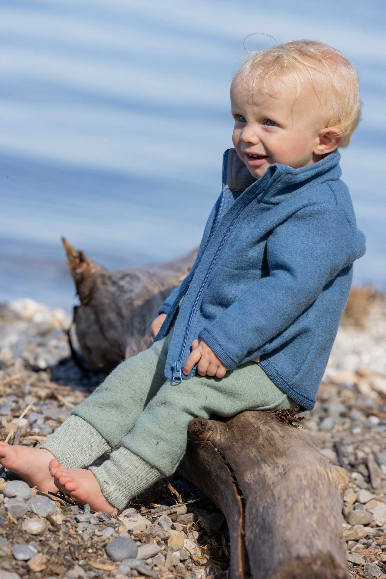 Blonder Junge in disana Zipper-Jacke, Taubenblau & Pumphose Jade, beide aus leichtem Walk sitzend auf Treibholz am Kiesstrand