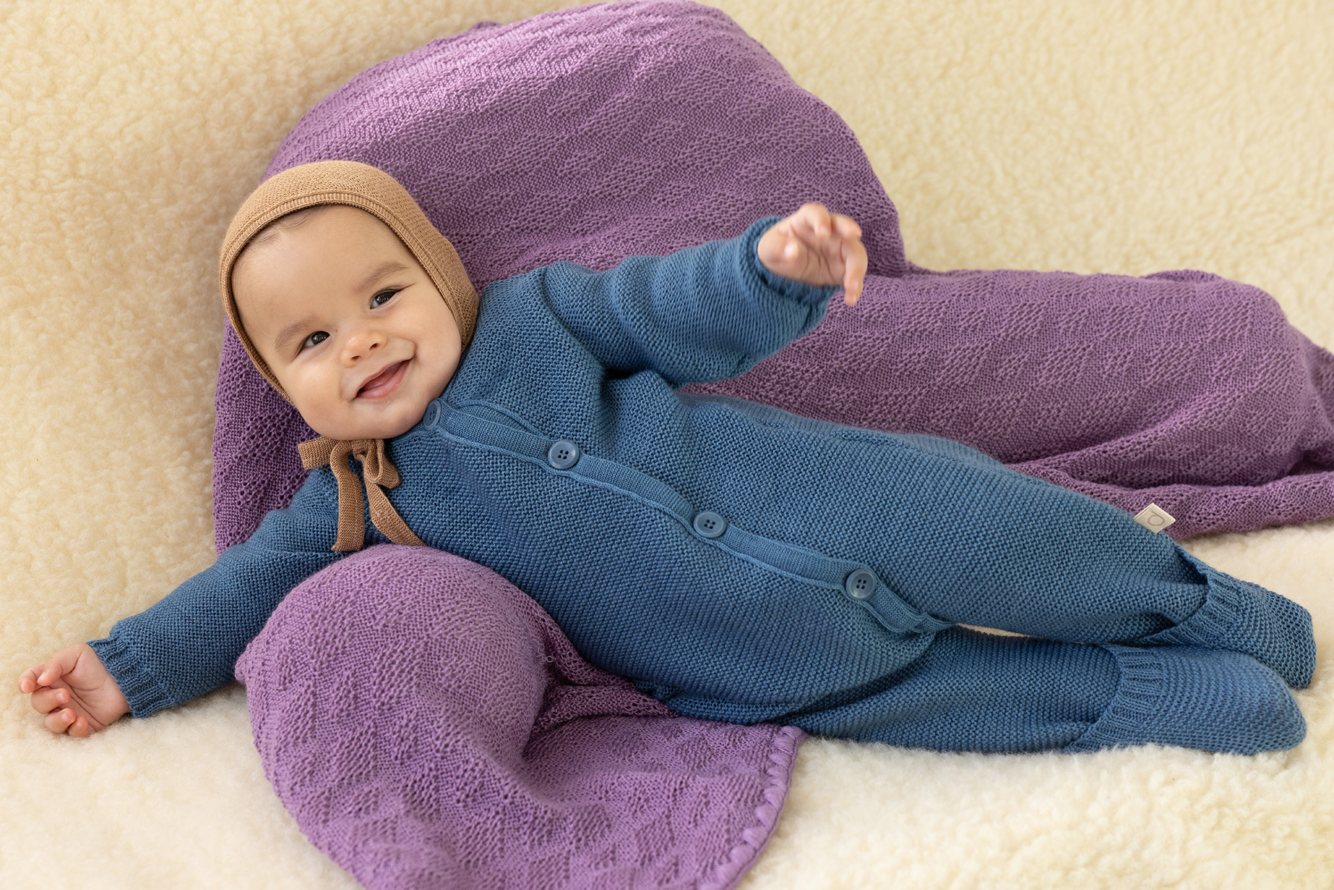 Lächelndes Baby in disana Strick-Overall, Taubenblau & einer Strick-Mütze in Karamell liegt auf einer Wolldecke in Flieder