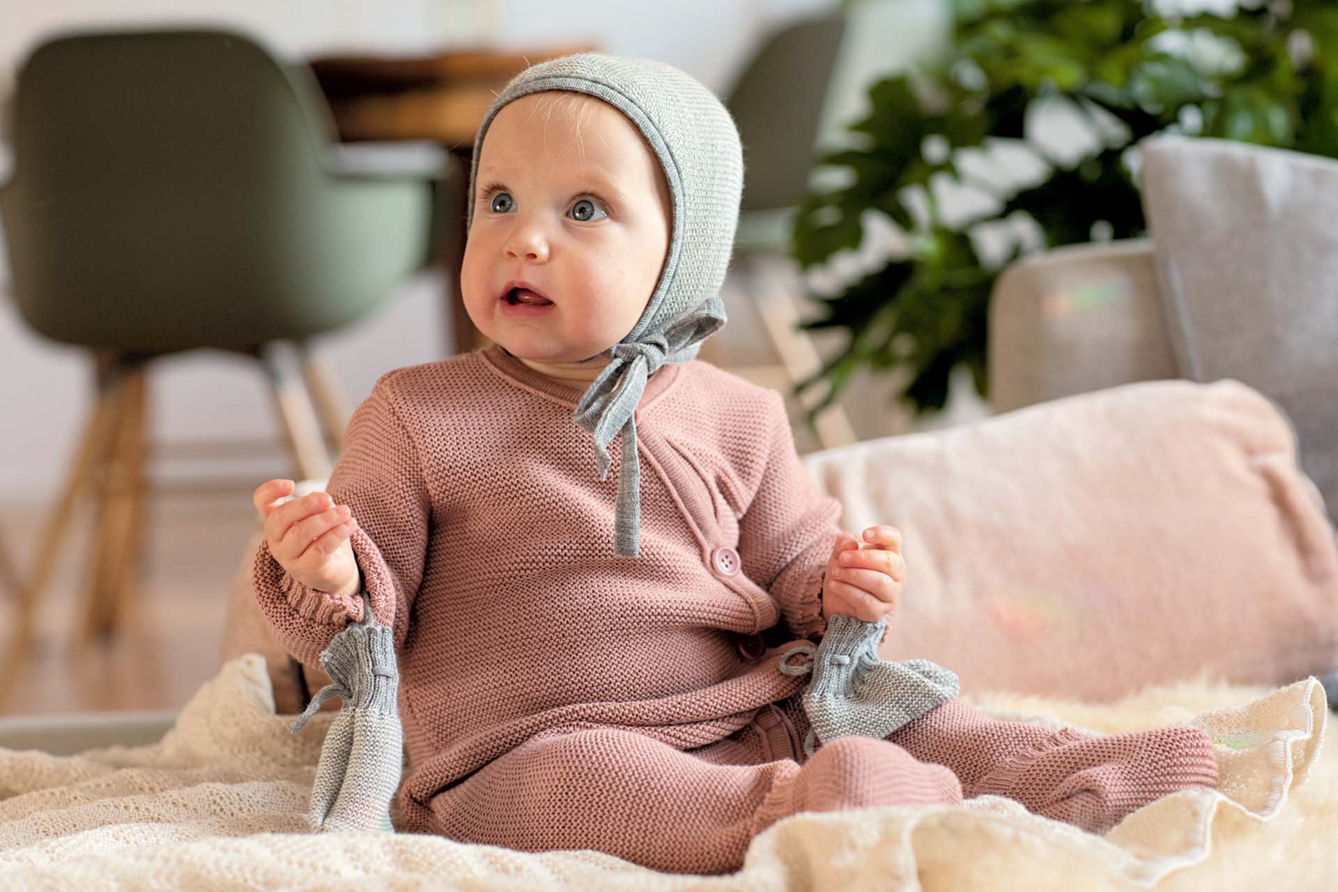Baby mit offenem Mund auf weicher Wolldecke Natur sitzend schaut nach oben links - bekleidet mit disana Strick-Overall Rosé