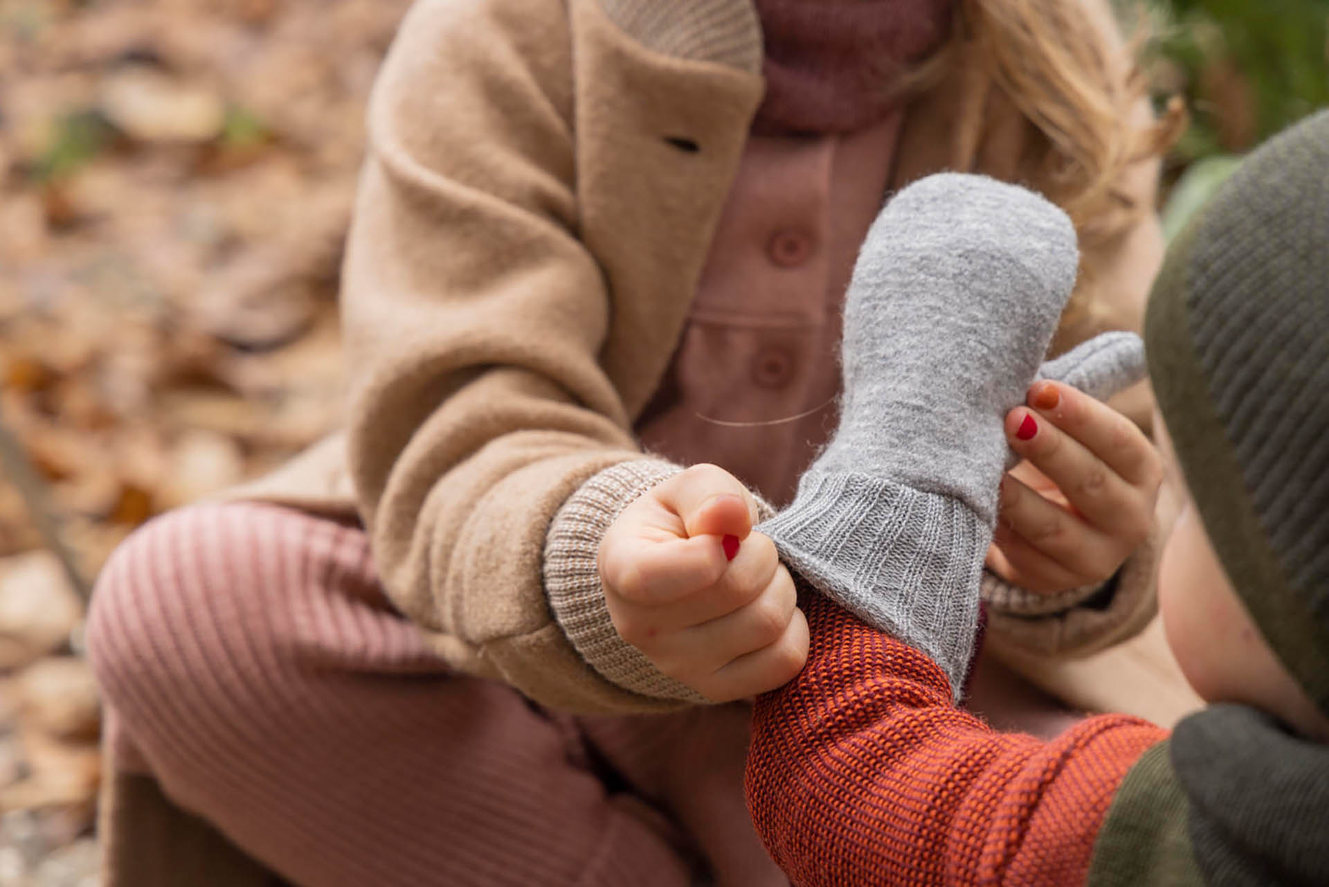 Mädchen im disana Walk-Mantel, Karamell, zieht einem Jungen eine disana Walk-Handschuh in Grau