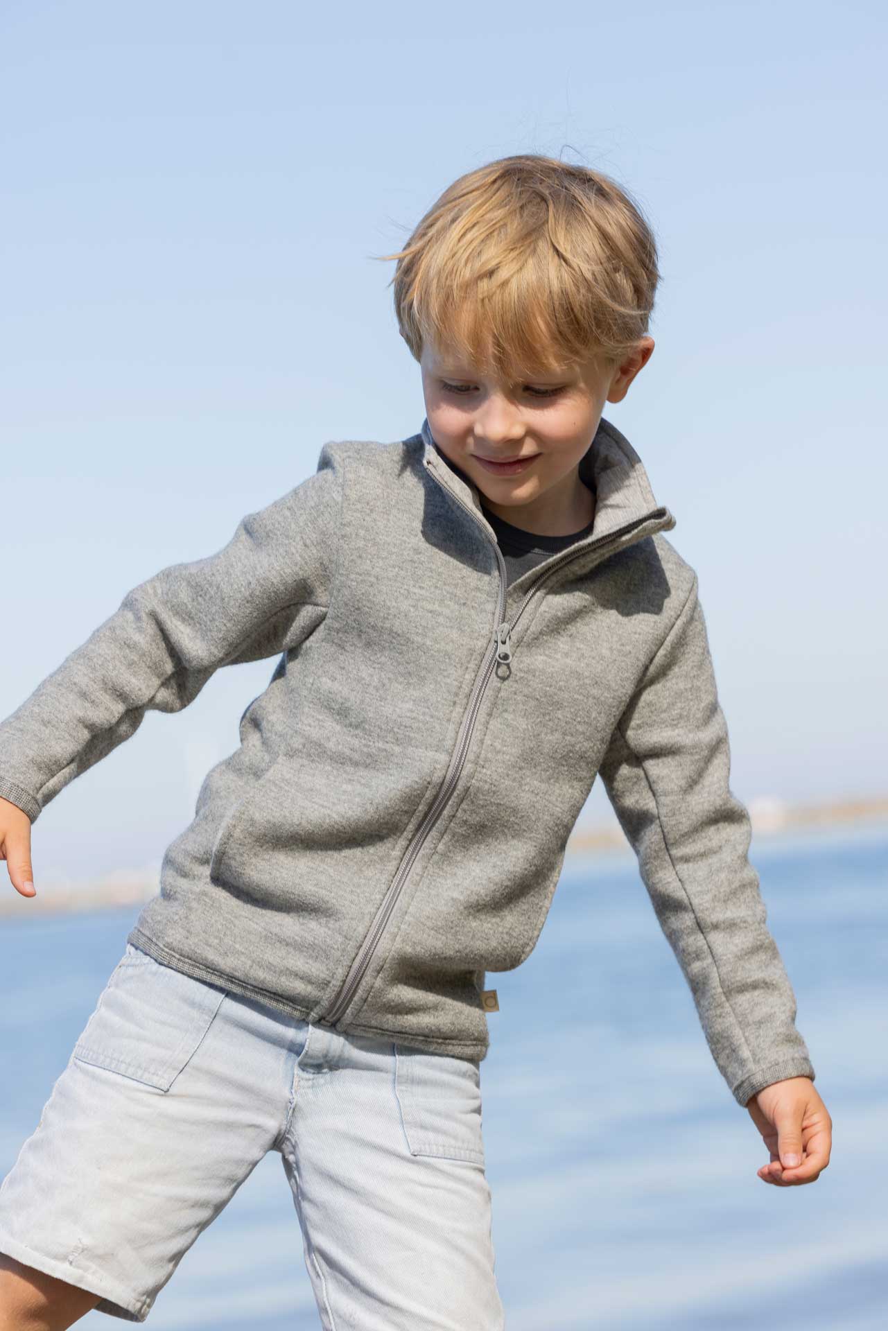 Blonder Junge in grauer disana Zipper-Jacke aus leichtem Wollwalk balanciert auf einem Bein am Seeufer.