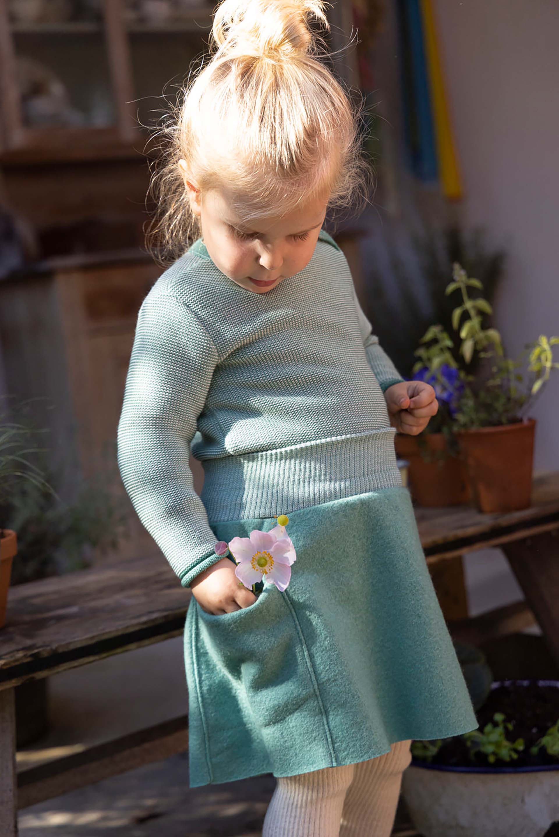 Blondes Mädchen im disana Melange-Pullover Mint-Natur und Walk-Rock in Mint steckt eine weiße Blume in Ihre Tasche