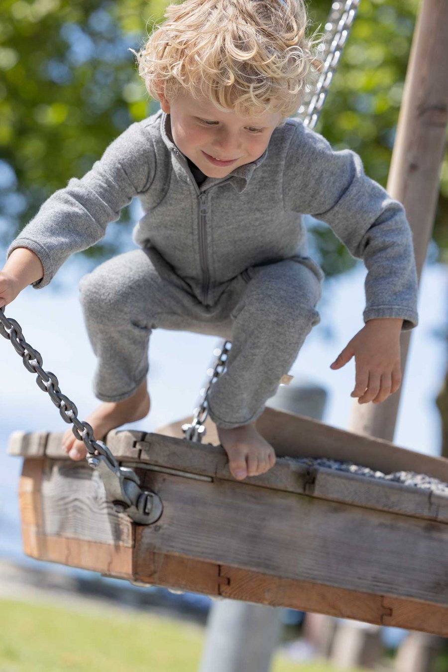 Kind im grauen disana Walkanzug auf Spielplatz am Seeufer, barfuß auf Schaukel, Sommer-Wollwalk 260g/m² aus Bio-Merinowolle
