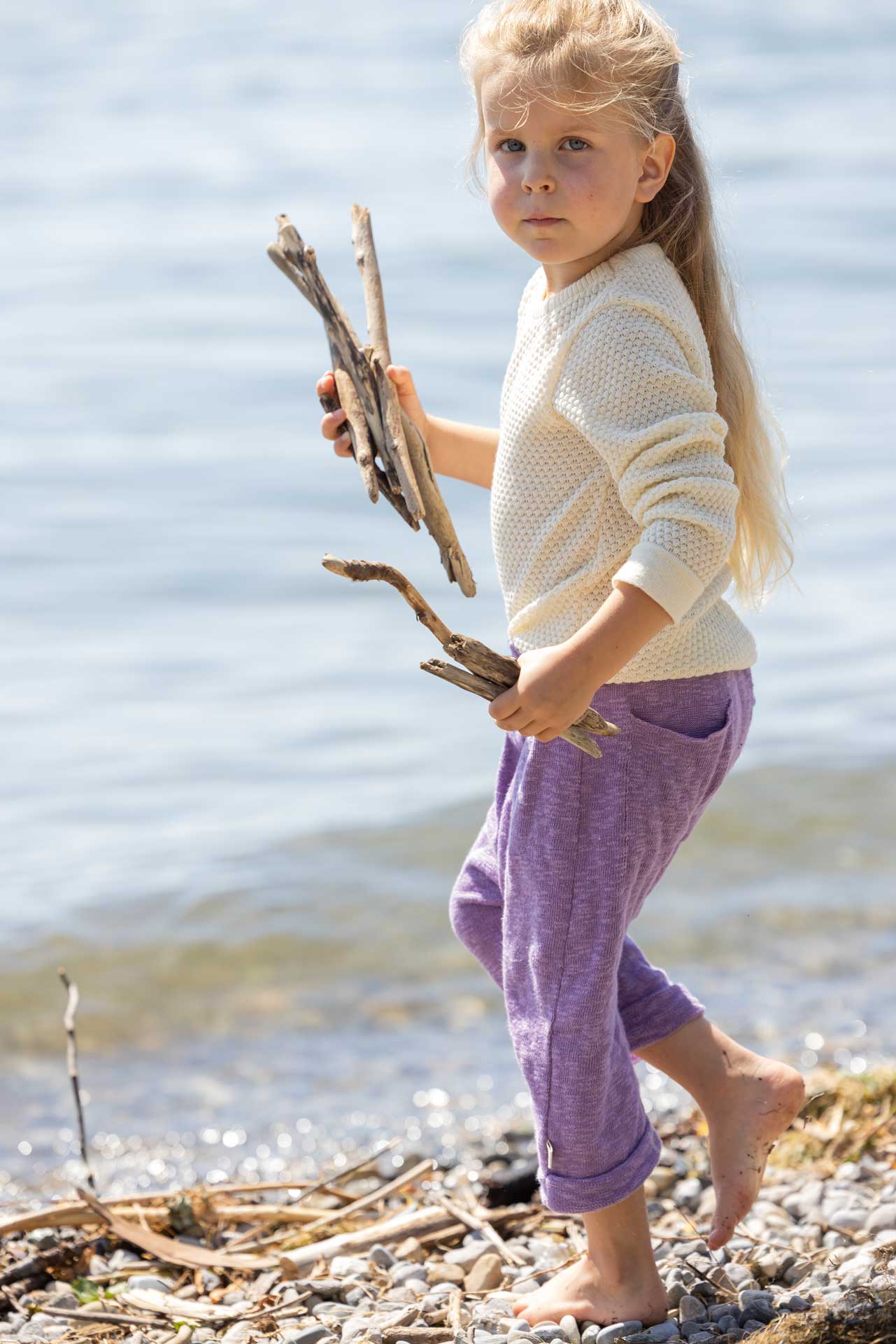 Junges blondes Mädchen in disana Wabenstrick-Pullover in Natur und Leinen-Strickhose in Flieder am Seeufer