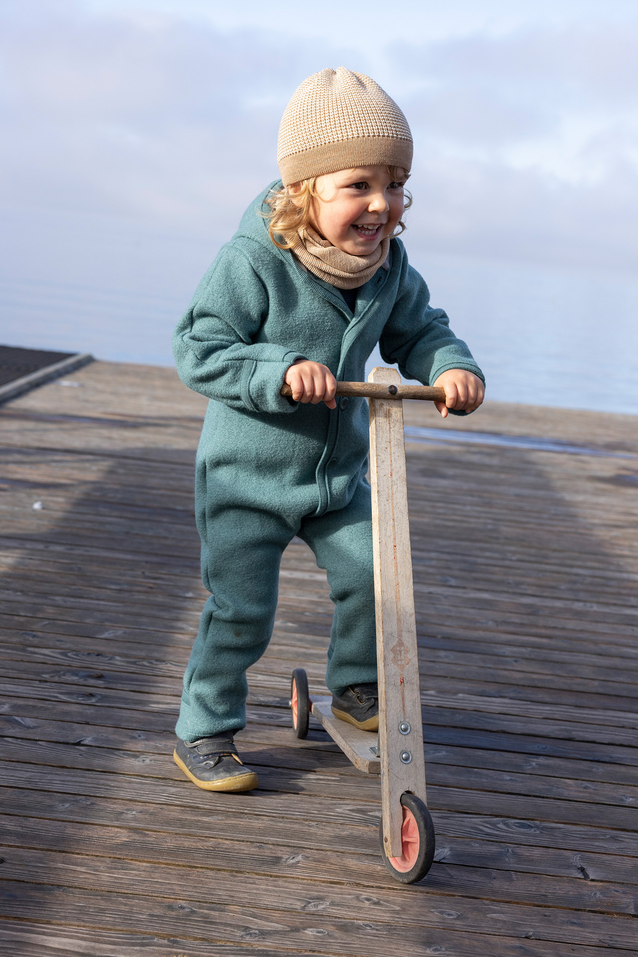 Lachendes blondes Kind in disana Walk-Overall Eukaylptus und Beanie Karamell-Natur fährt Holzroller auf einem Steg am See.