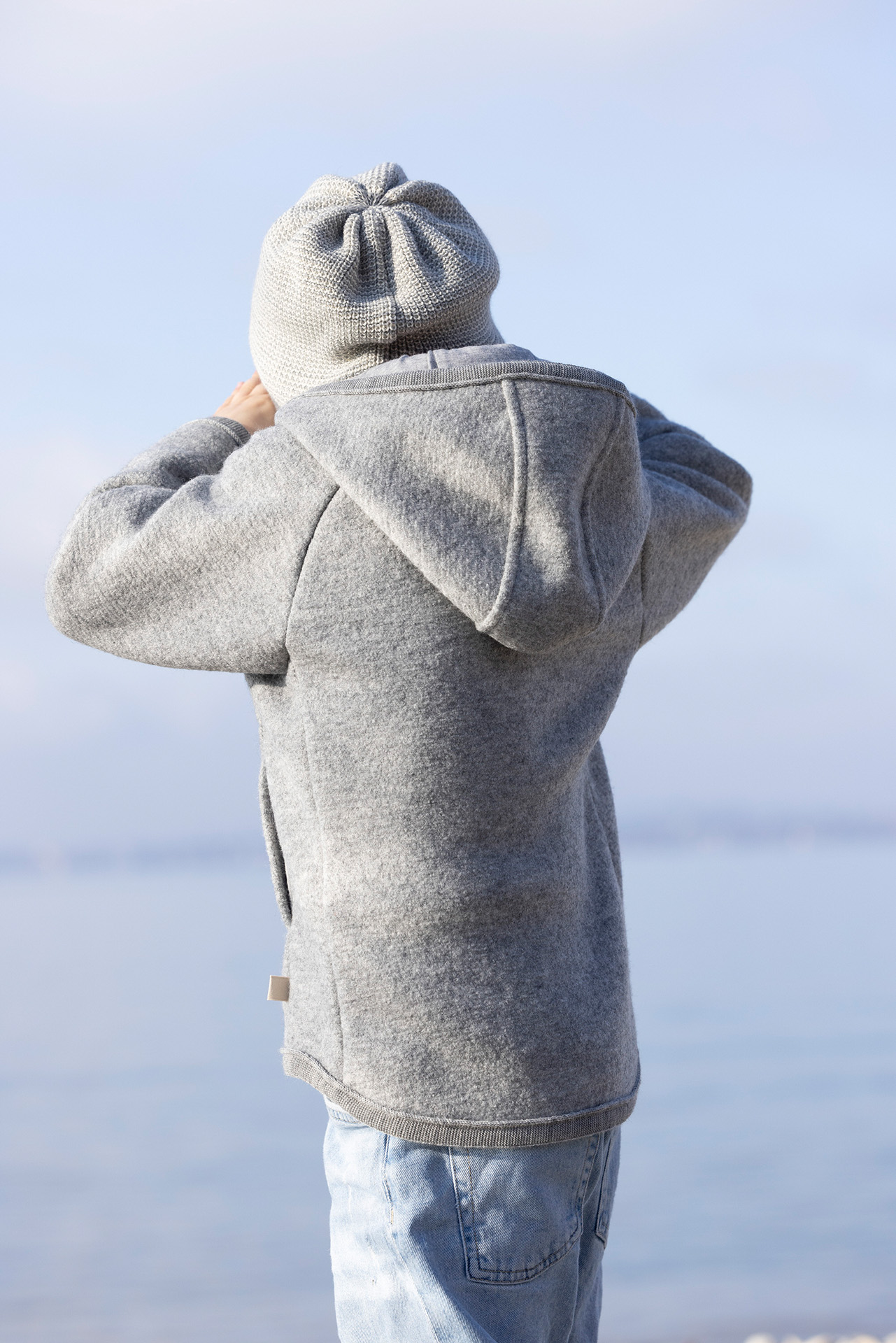 Rückansicht: Kind in grauer disana Walk-Jacke Zipp mit Kapuze und Beanie in Grau-Natur & Jeans blickt auf den See