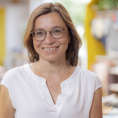 Elisabeth Courbet, Verwaltungsangestellte bei disana in weißer Bluse vor unscharfem Hintergrund lächelt in die Kamera.