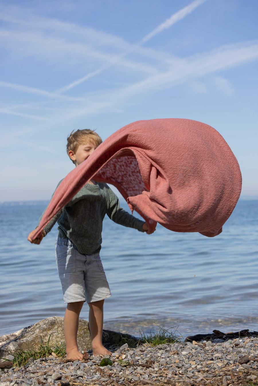 Ein blonder Junge spielt am Seeufer mit einer disana Leinen-Decke in Softpink. Der Wind bläst die Decke auf.