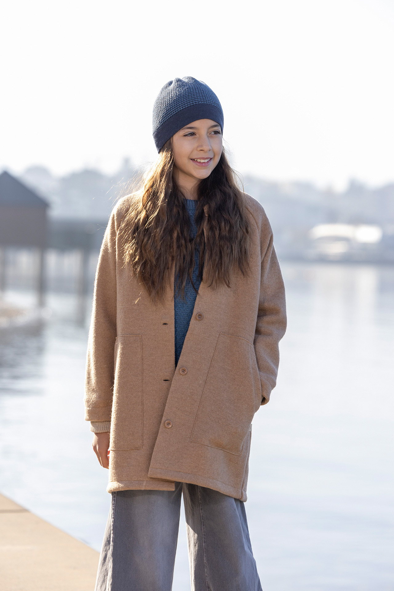 Braunhaariges Mädchen im Teenager-Alter mit disana Walk-Mantel Karamell und Beanie in Indigo-Taubenblau steht am Seeufer