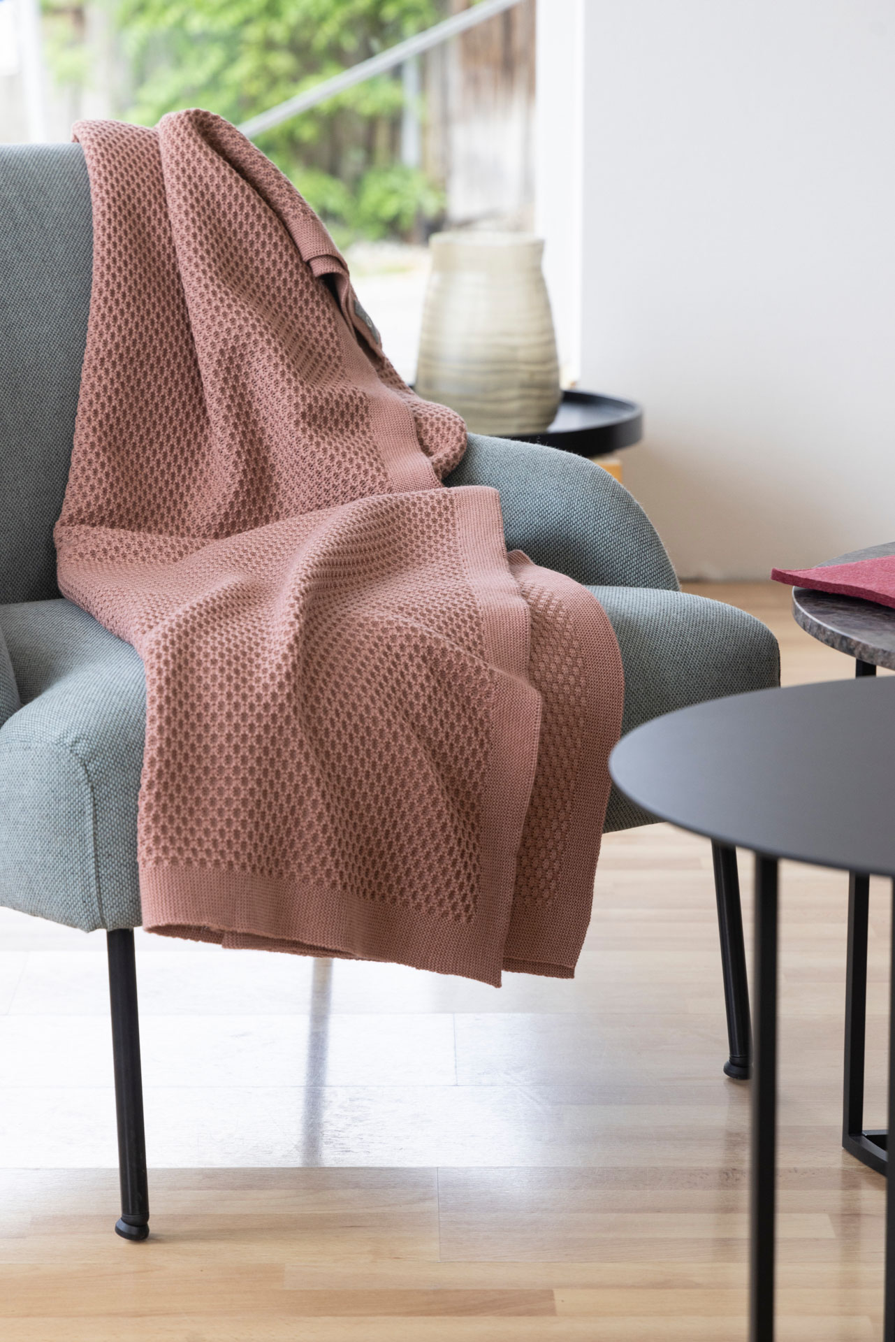 disana Waben-Strickdecke, Rosé auf einem hellgrauen Sessel drapiert in einem hellen Wohnzimmer mit Holzparkett & Tischchen