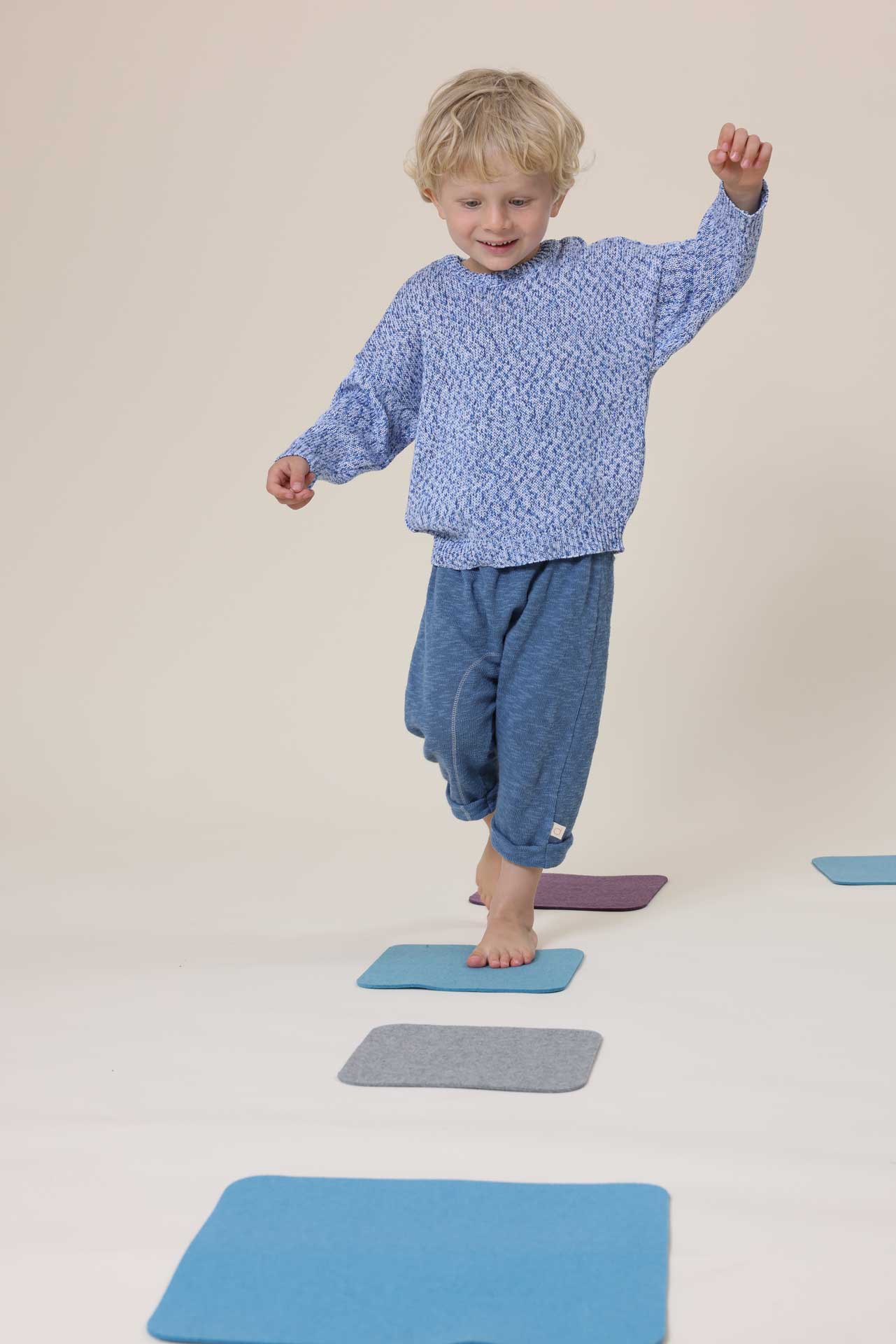 Kind in blauem disana Baumwoll-Pullover "Spray" und Leinen-Strickhose spielt barfuß auf bunten Filzmatten aus Recycling-Filz