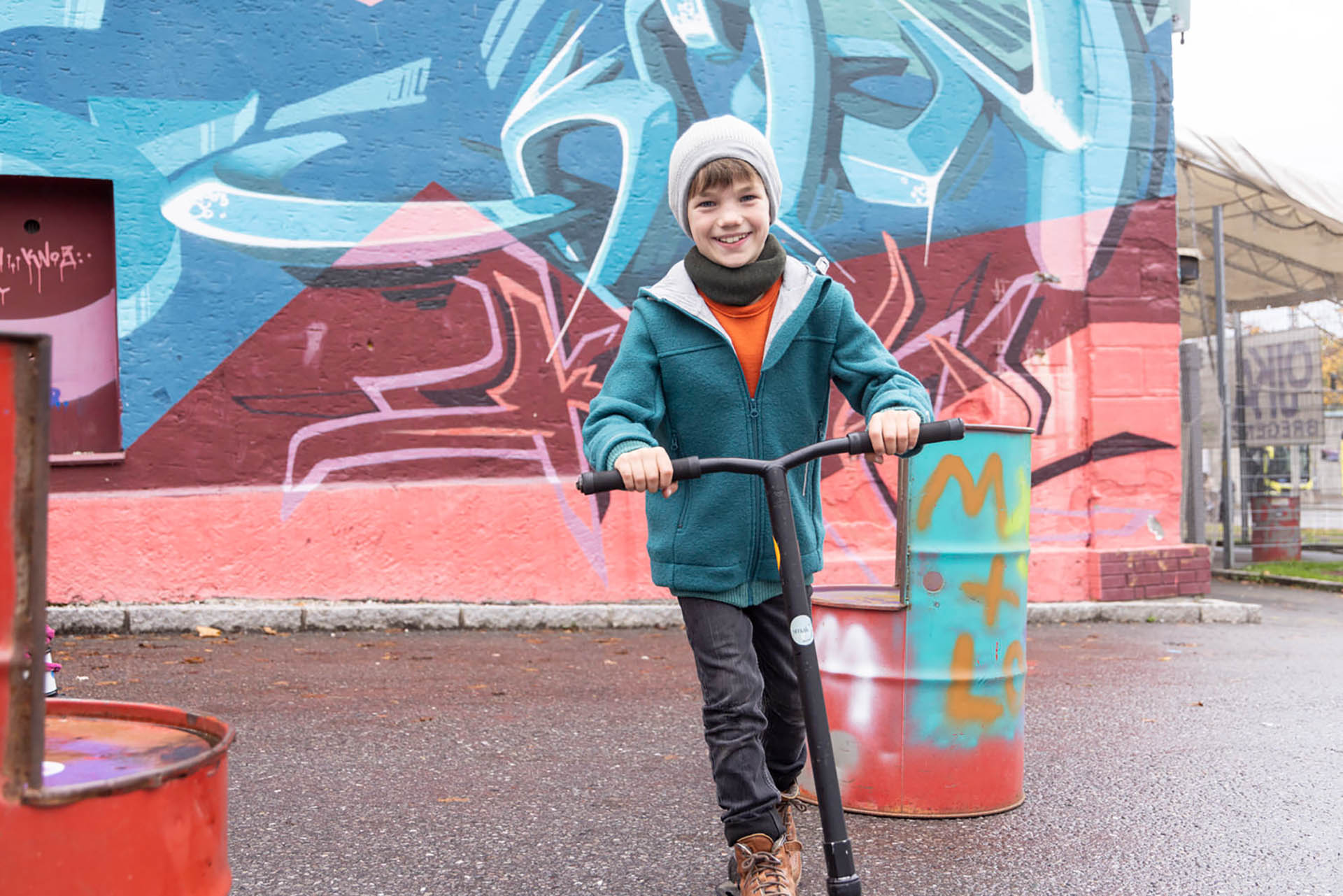 Ein Junge in disana Outdoor-Jacke in Pacific und Beanie Grau-Natur lacht auf einem Roller vor einer bunten Graffiti-Wand.