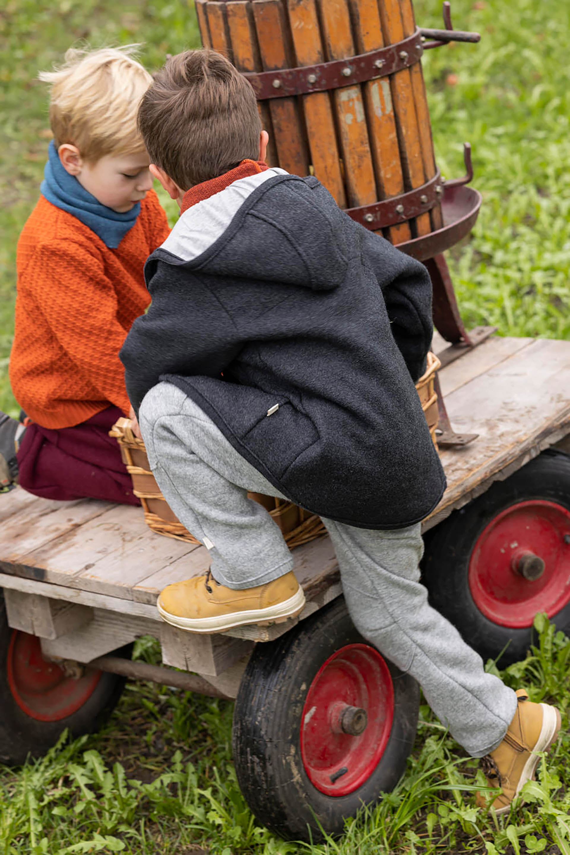 Zwei Jungen in disana Kleidung aus Wollwalk spielen auf einem Holzwagen mit einer alten Obstpresse und Weidenkörben im Garten