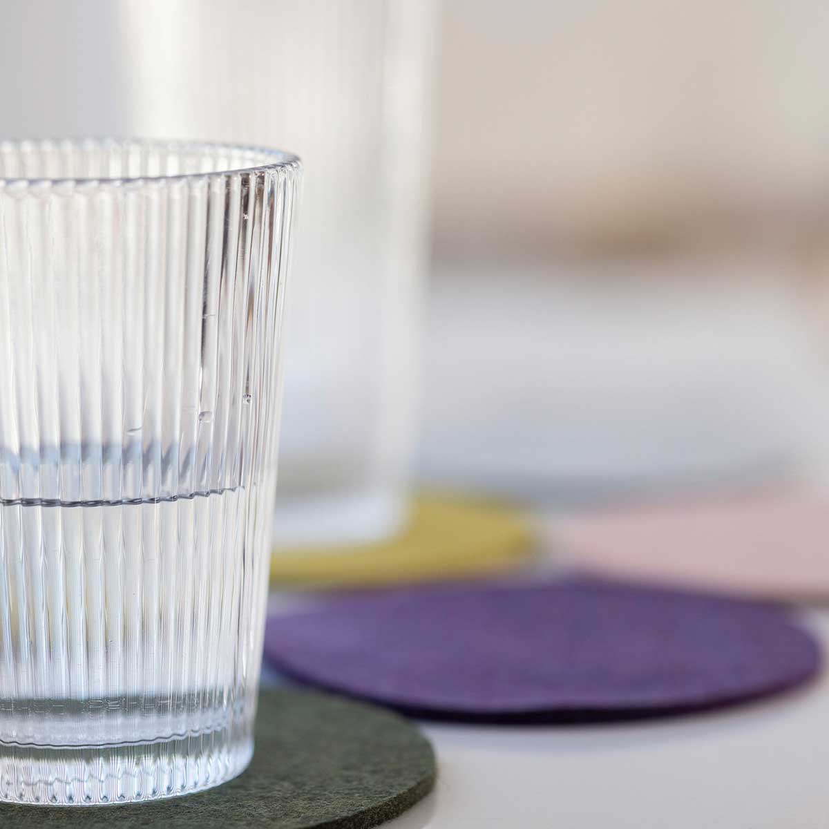 Halbvoller Riffelglas auf olivem disana Filz-Untersetzer; weitere bunte Wollfilz-Untersetzer unscharf im Hintergrund.