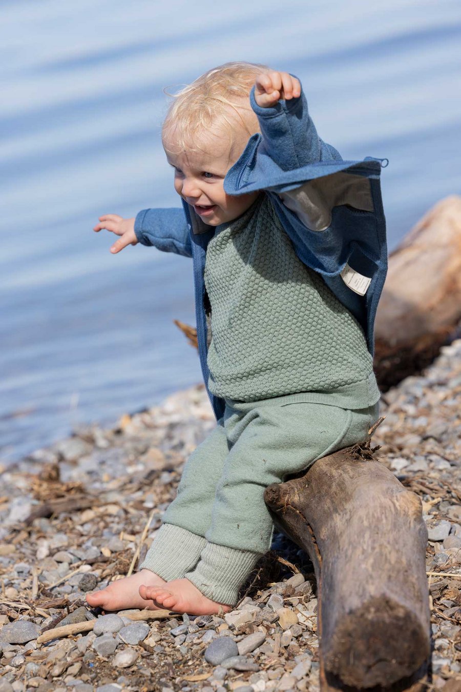 Blondes Kind in disana Zippper-Jacke Taubenblau, Pumphose & Wabenstrick-Pullover in Jade am Wasserufer auf Baumstamm sitzend
