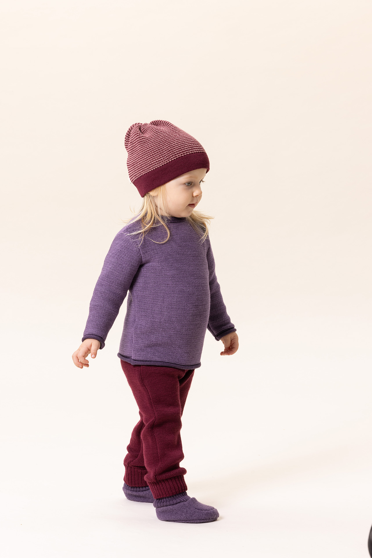 Blondes Kind in disana Melange-Pullover Aubergine-Flieder, Strick-Trägerhose Cassis Beanie Cassis-Rosé vor hellem Hintergrund