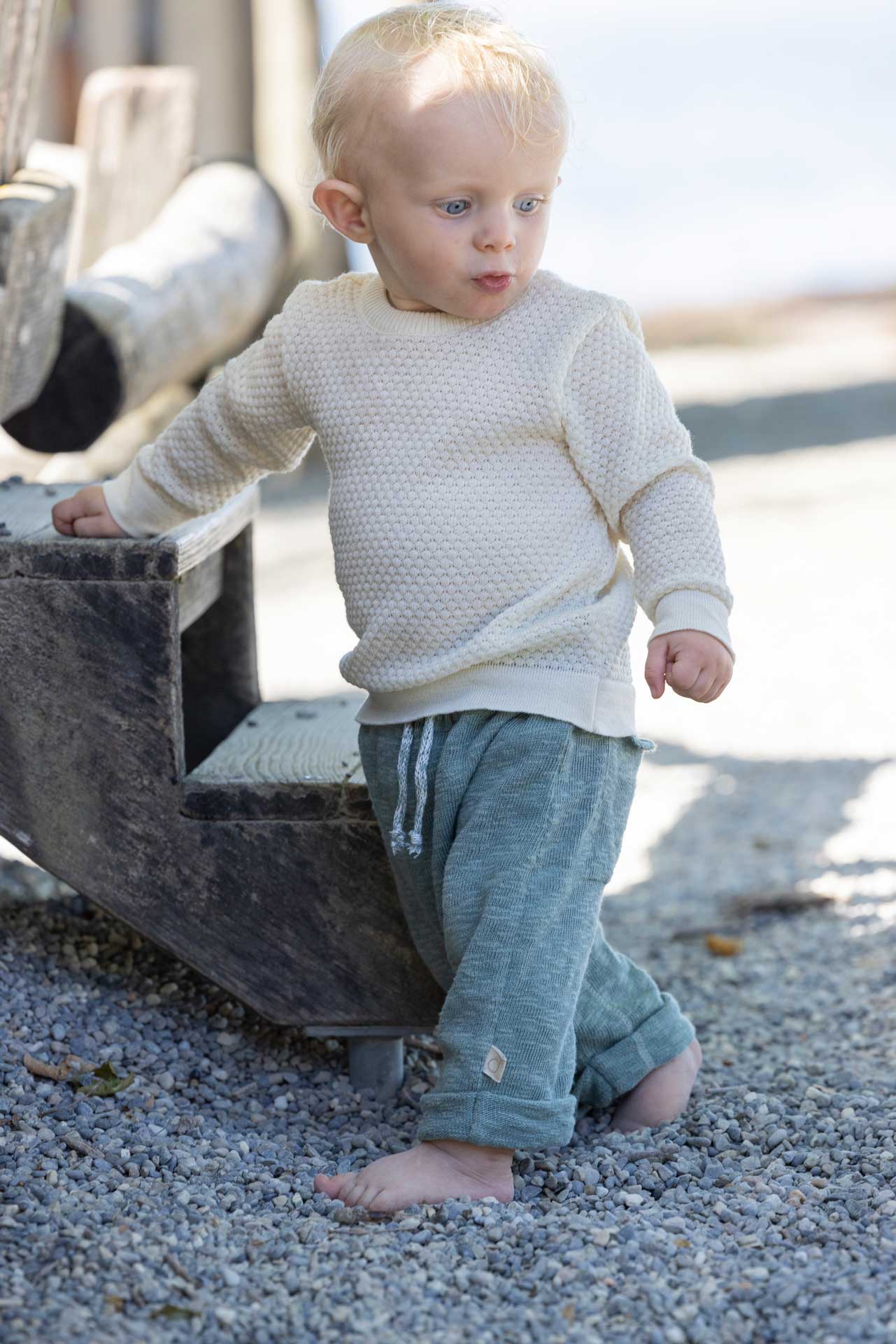 Blondes Kind in disana Wabenstrick-Pullover in Natur und Leinen-Strickhose in Jade lehnt an einer verwitterten Holztreppe
